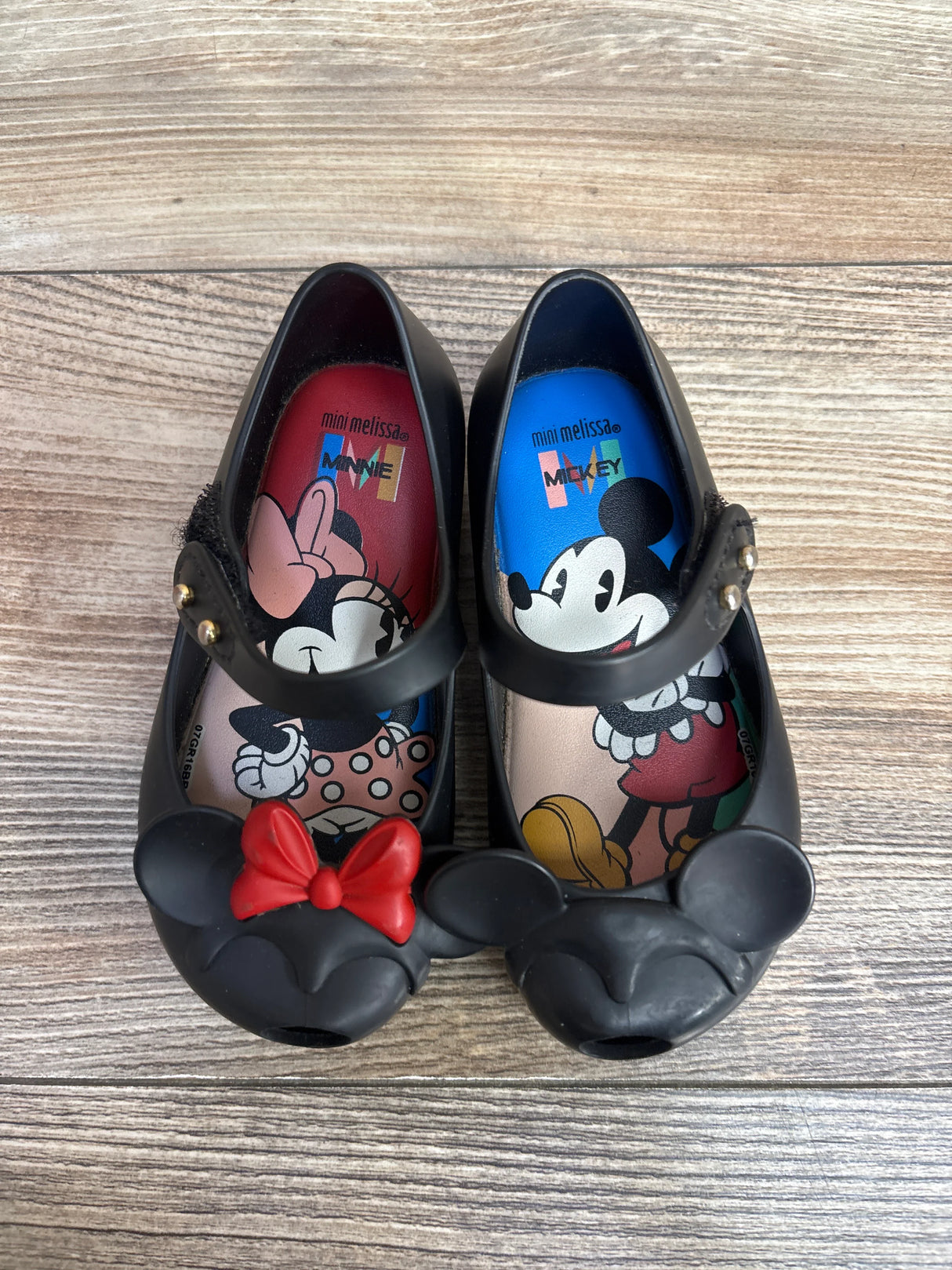 Black toddler shoes featuring Minnie and Mickey Mouse designs, with Minnie’s red bow detail, on a wooden floor
