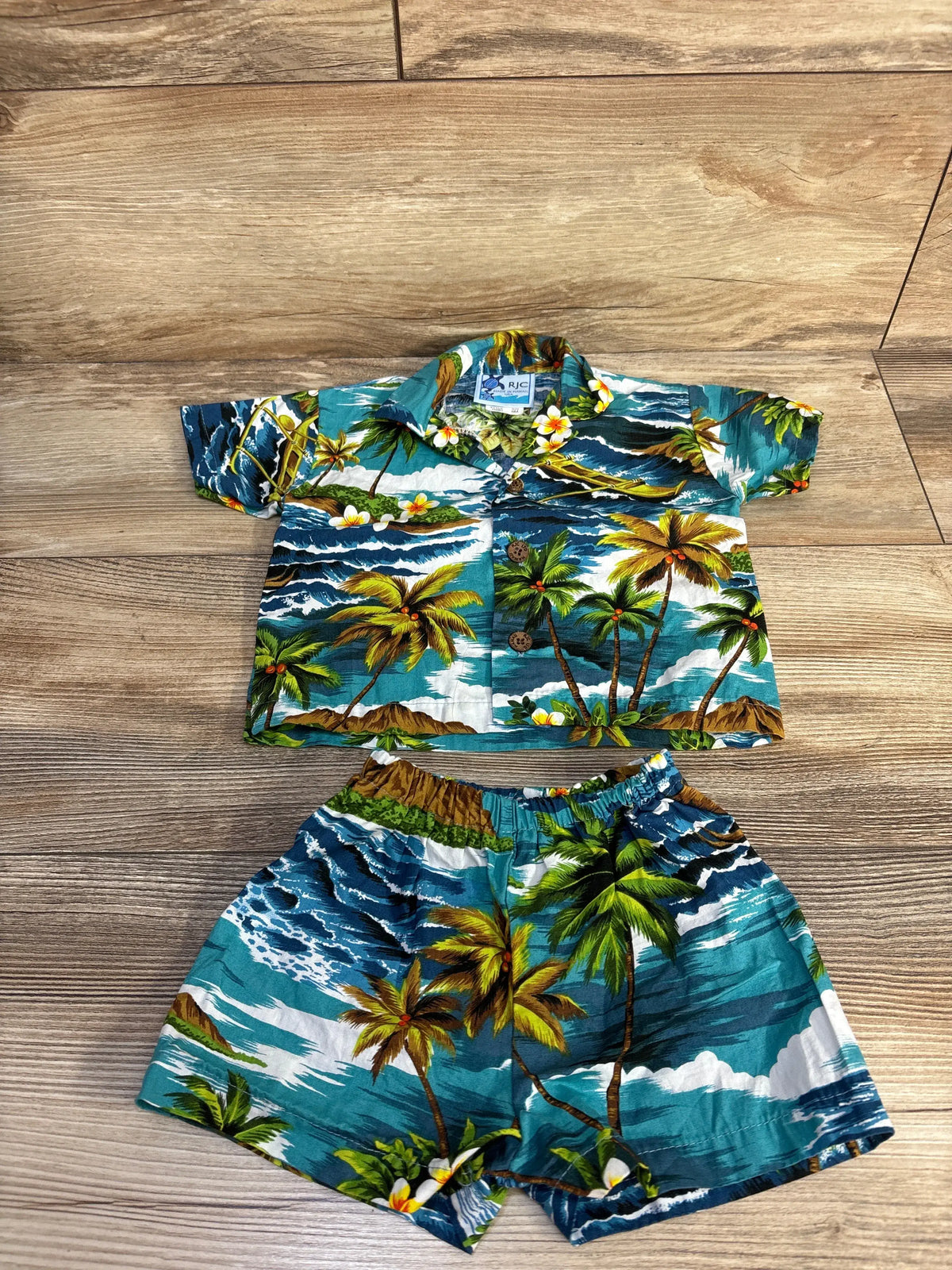 Flat lay of blue RJC two-piece button-up shirt and shorts set showcasing tropical palm print with a wood background