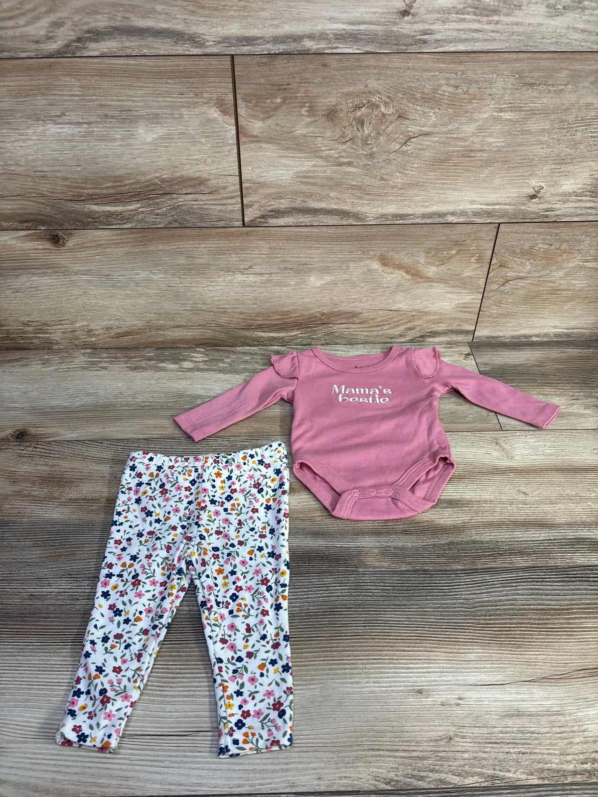 Flat lay of pink long-sleeve bodysuit and white floral pants showing Mama’s bestie text and ruffle shoulders on wood
