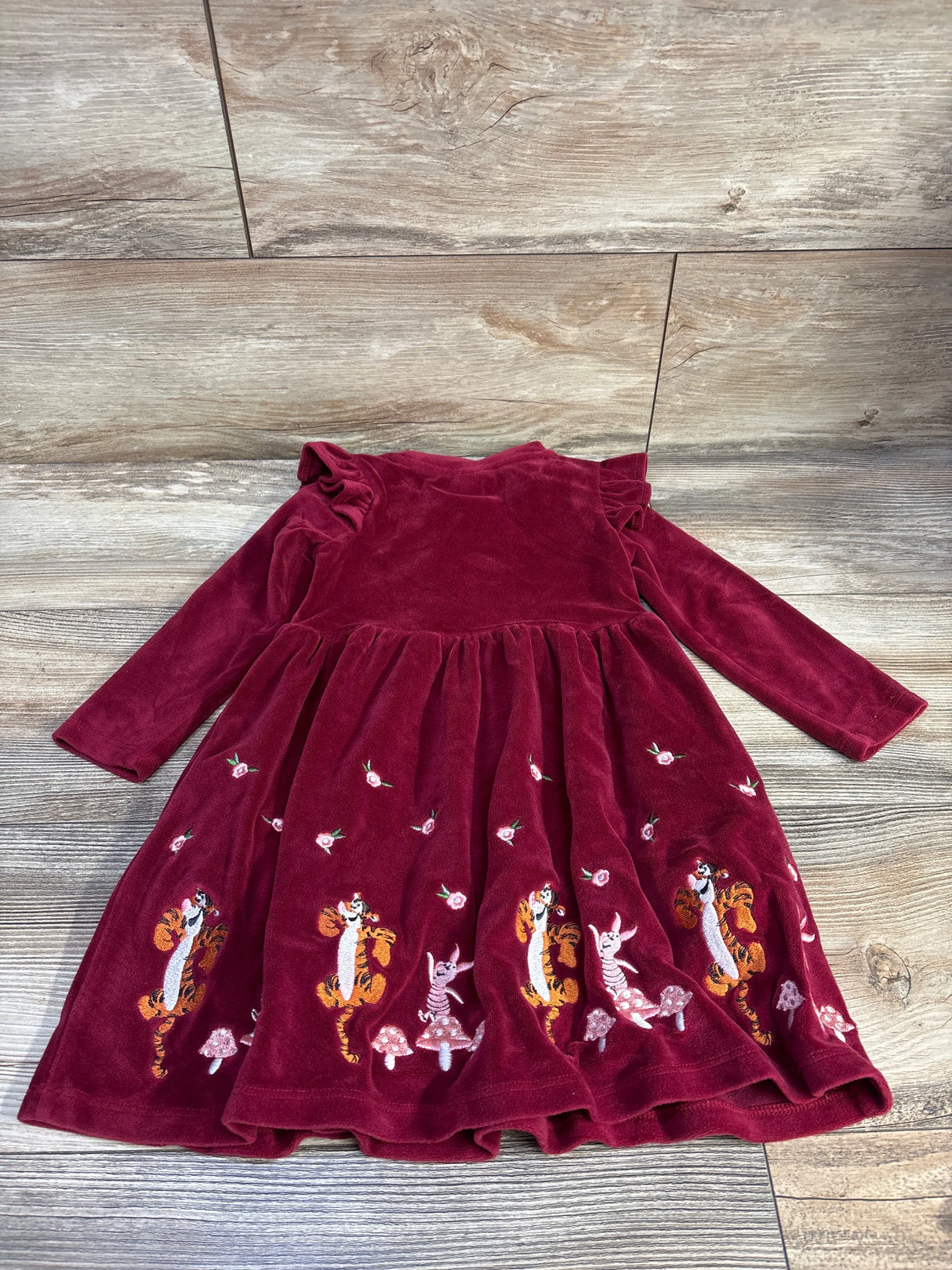 Flat-lay of maroon velvet Winnie the Pooh long-sleeve toddler dress showing embroidered Tigger and Piglet hem and ruffled shoulders on wood background