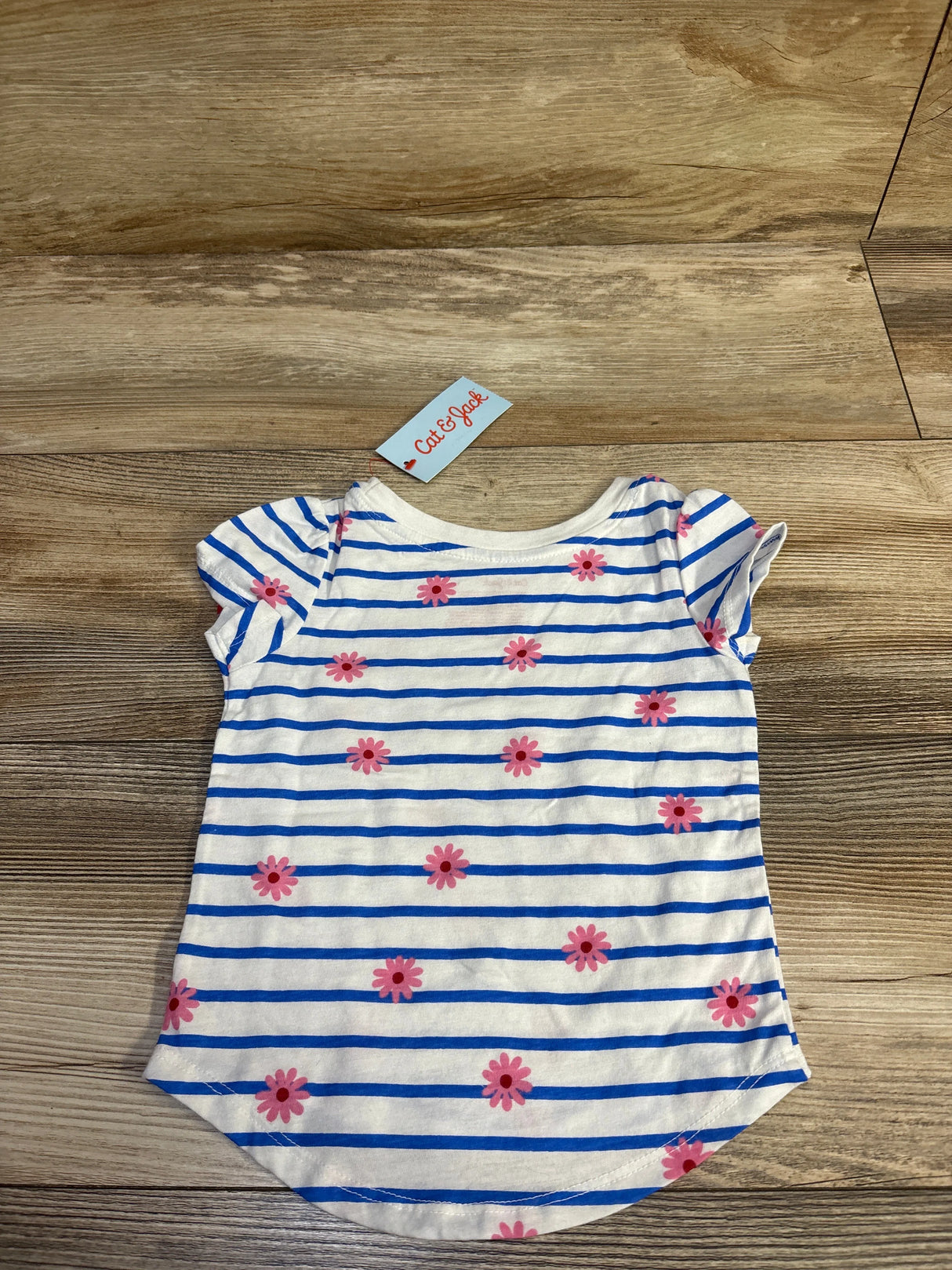 Front flat lay of Cat & Jack baby shirt showcasing blue horizontal stripes and pink daisy print with short cap sleeves on wood background