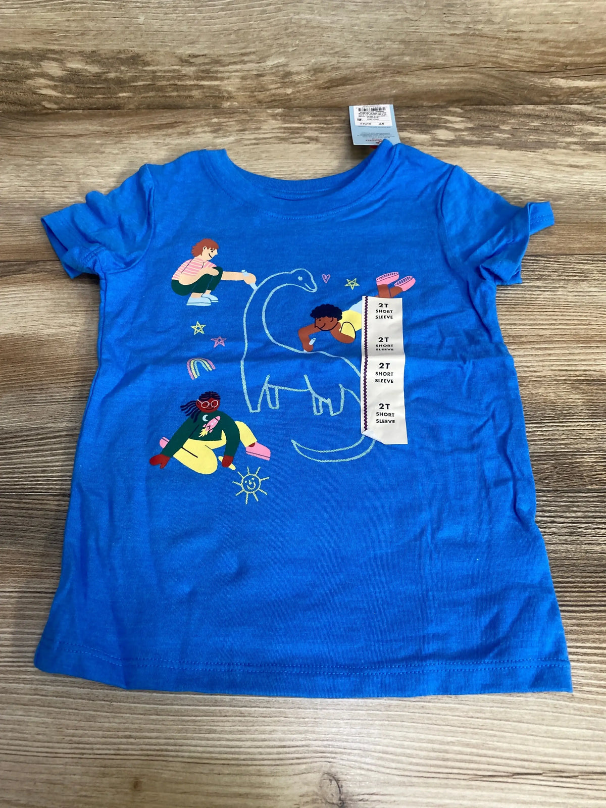 Flat lay of blue Cat and Jack 2T short-sleeve shirt showing chalk-style dinosaur and kids graphic on wood background