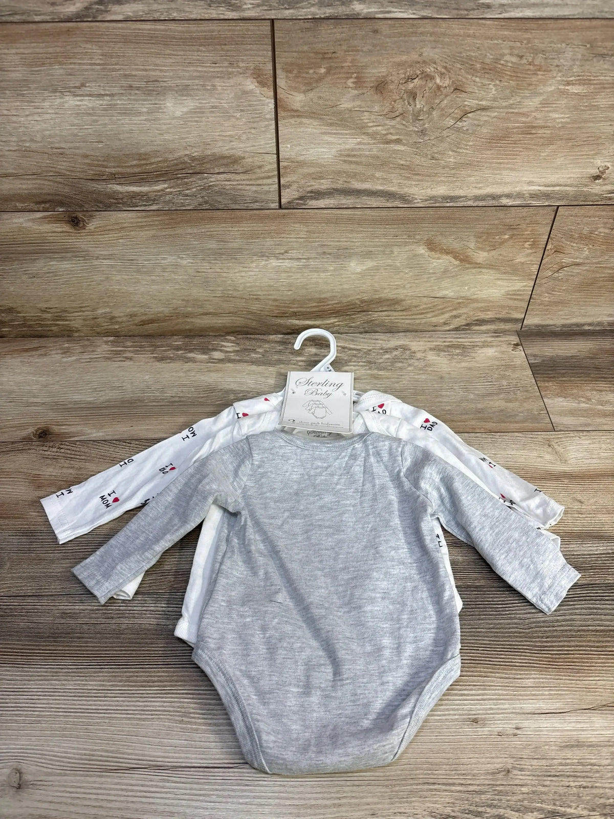 Front view of 3-pack Sterling Baby long sleeve bodysuits, grey top and two white printed, against wood floor