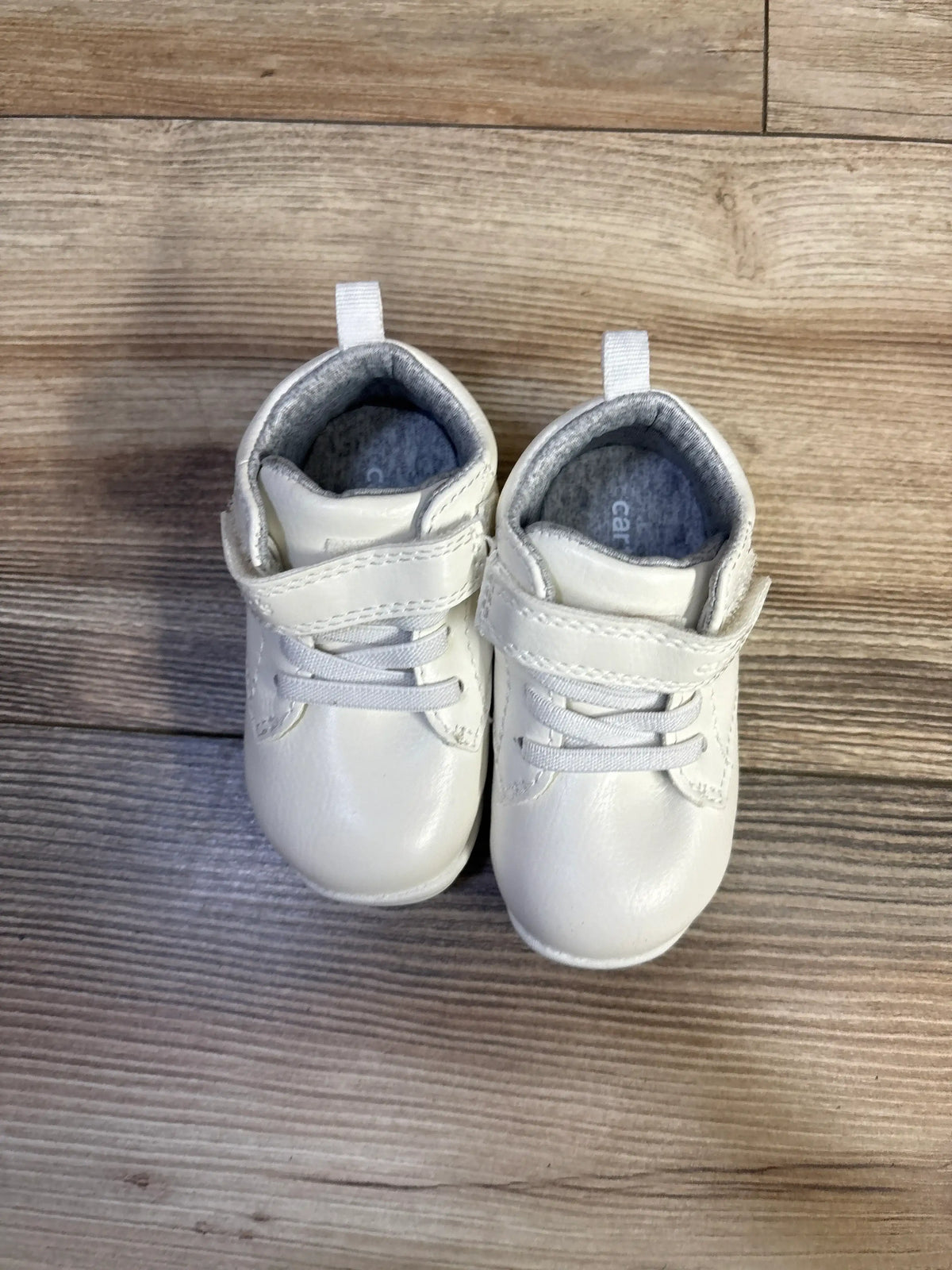 Top-down view of white Carter’s high-top baby sneakers showing Velcro strap, elastic laces and grey lining on wood floor