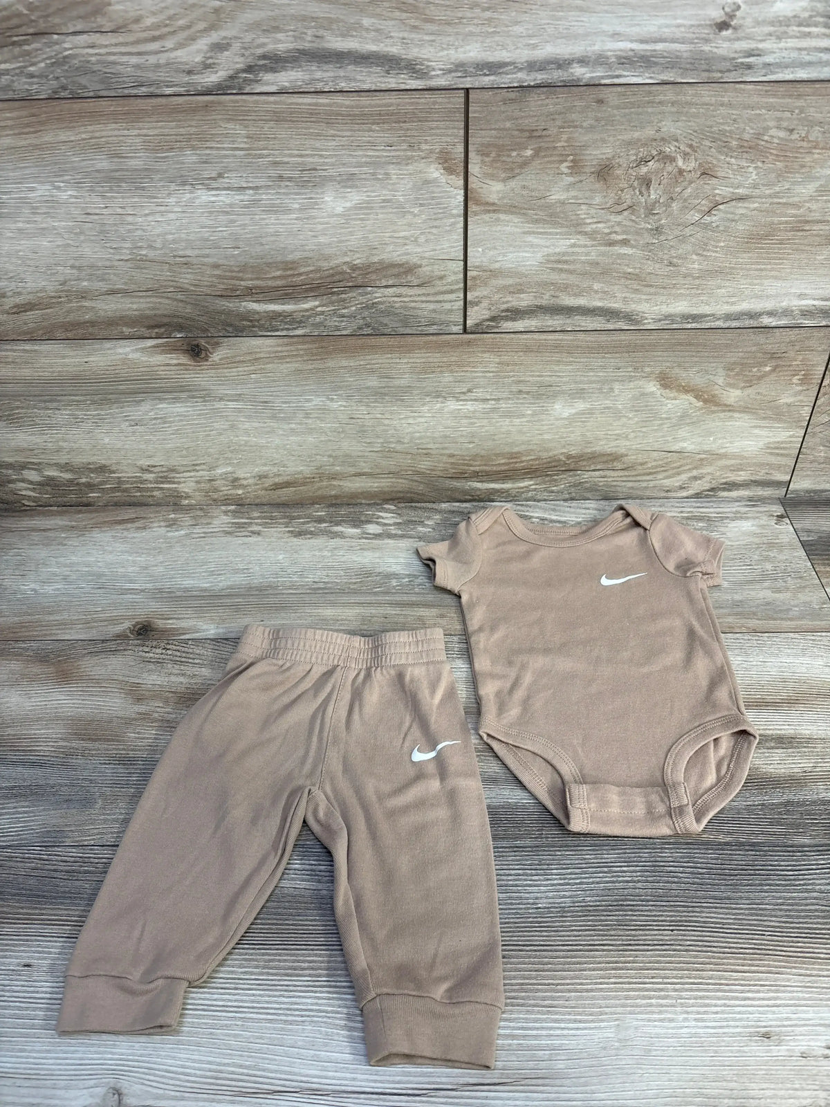 Flat lay of tan Nike 2-piece baby bodysuit and pants, size 6M, showing white Swoosh logos, elastic waistband and snap closures on light wood background