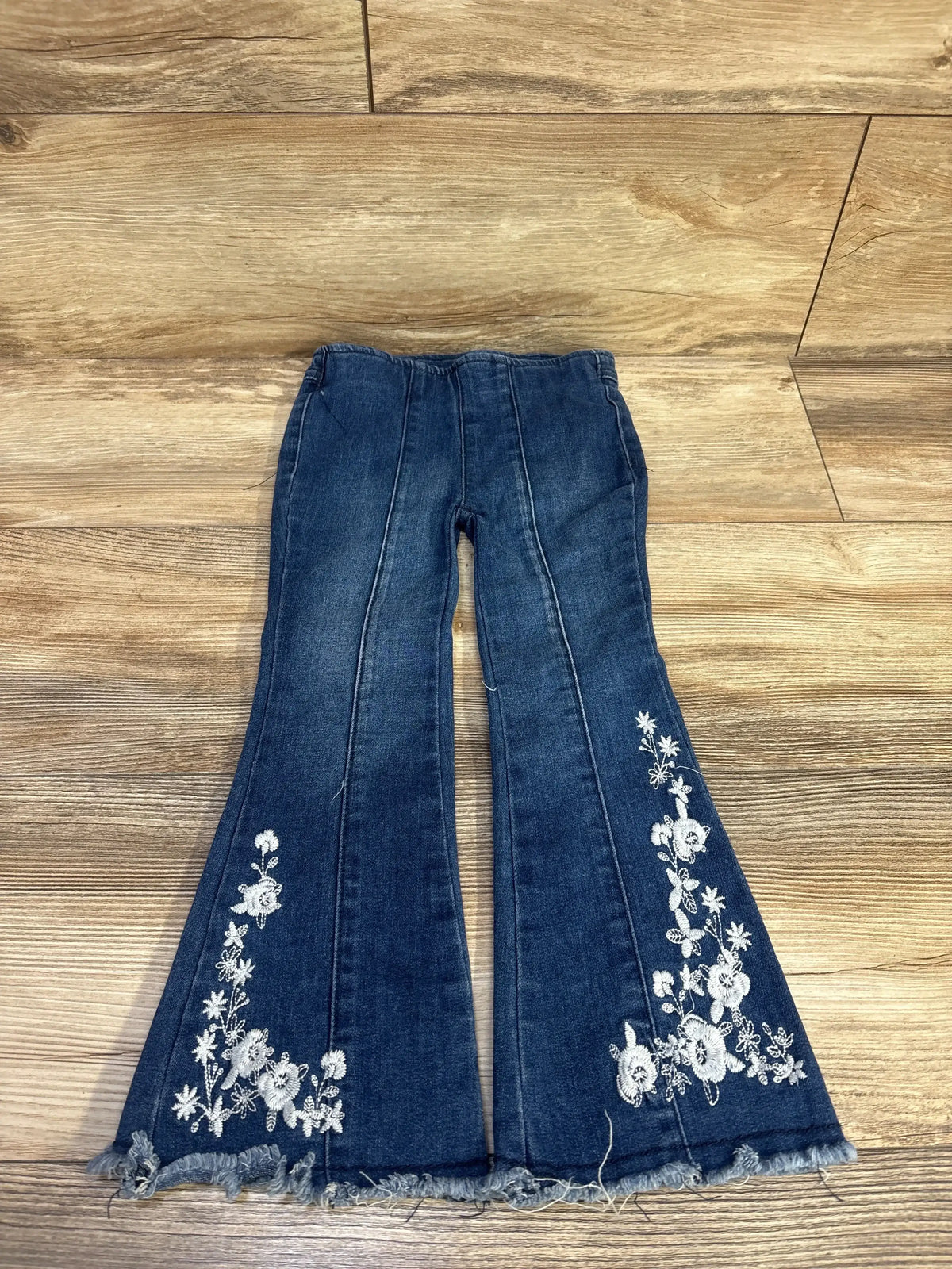 Flat lay of blue toddler flare jeans size 2T with white floral embroidery on lower legs and frayed hems on wood background