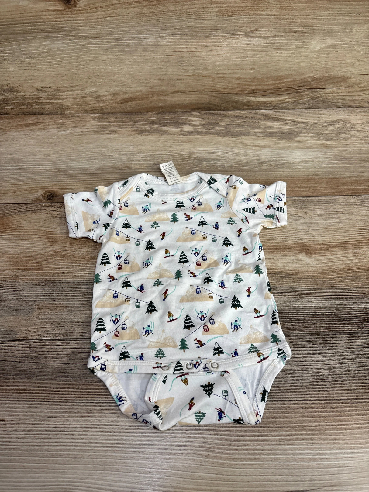 White baby onesie with ski-themed print of skiers, trees, and ski lifts on wooden background
