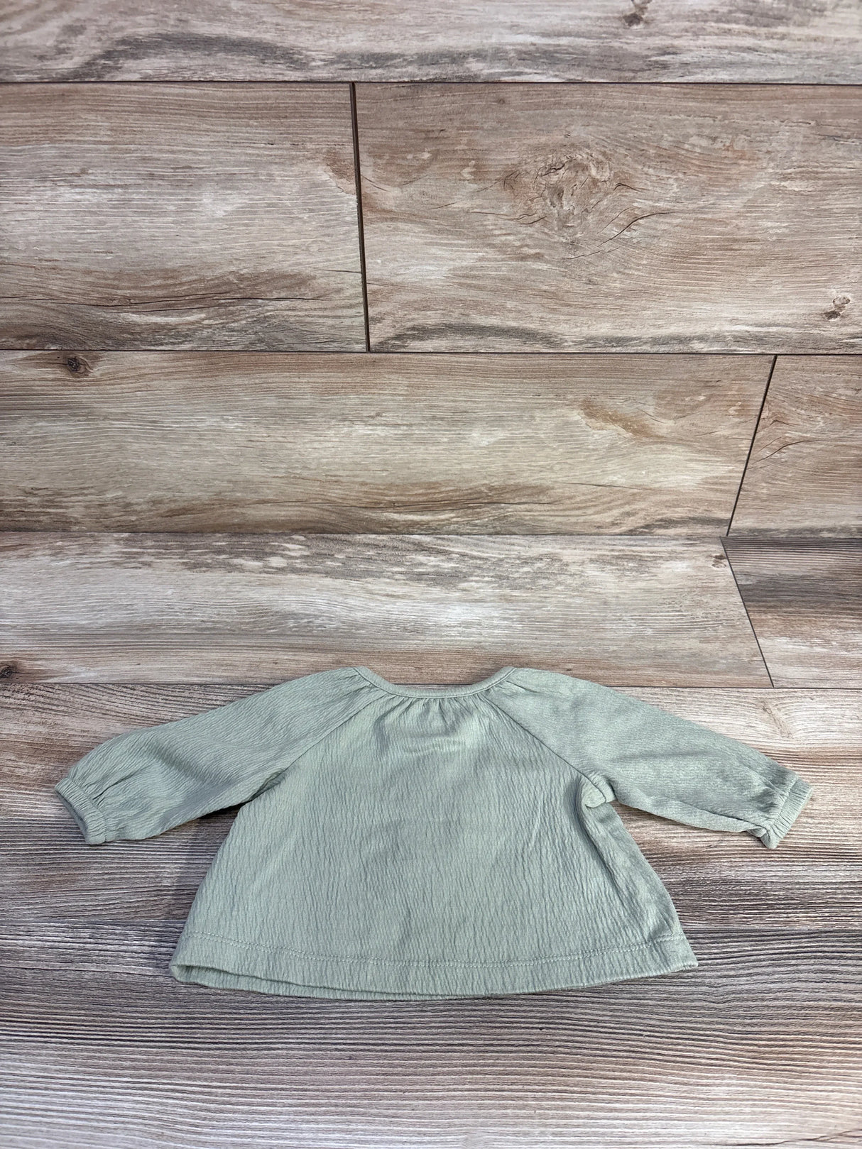 Flat lay of Carter's long-sleeve henley baby shirt showing back with gathered neckline, sage green cotton on wood plank background