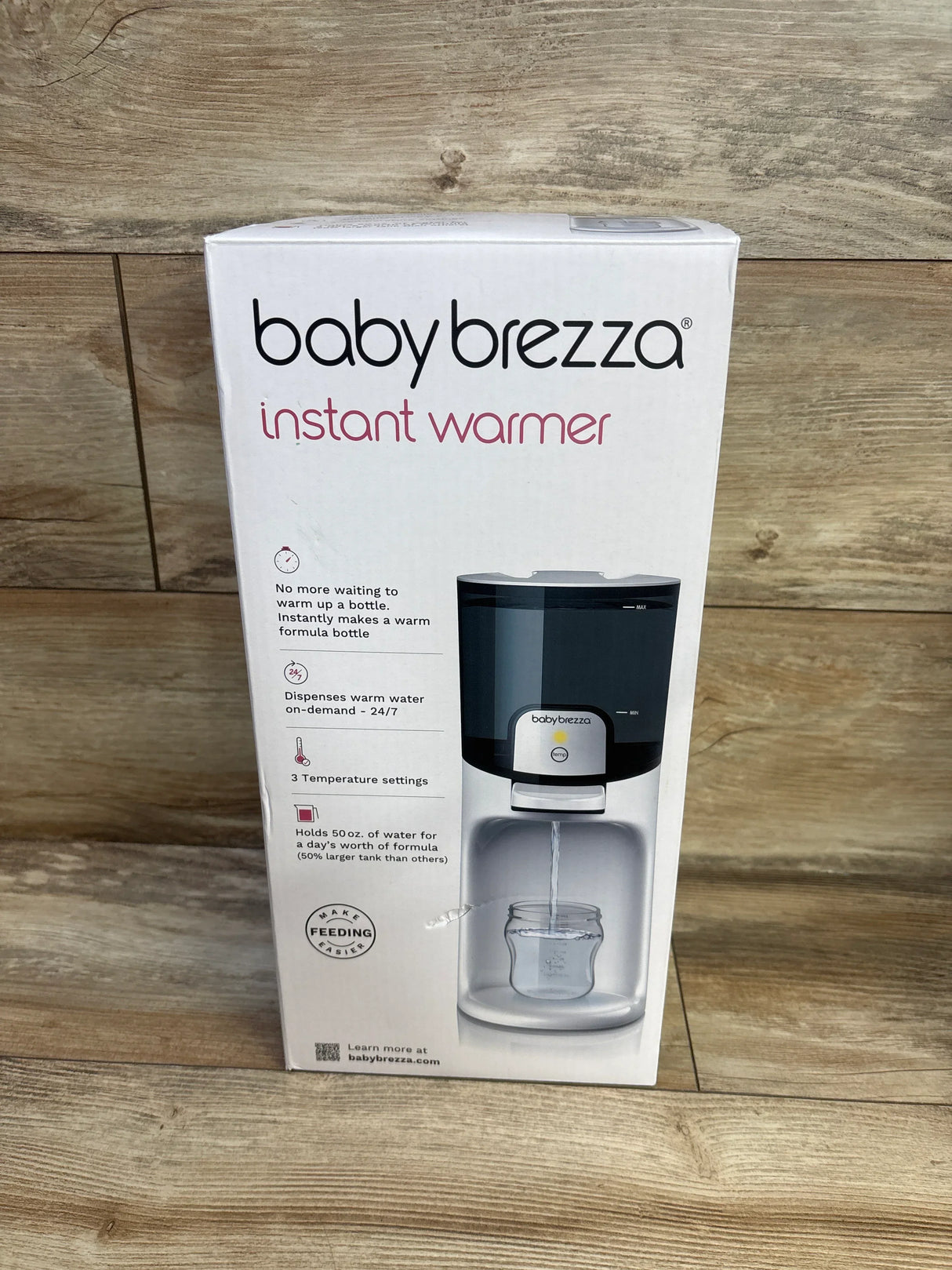 Front view of Baby Brezza Instant Warmer box showing the unit dispensing warm water into a baby bottle on wood floor