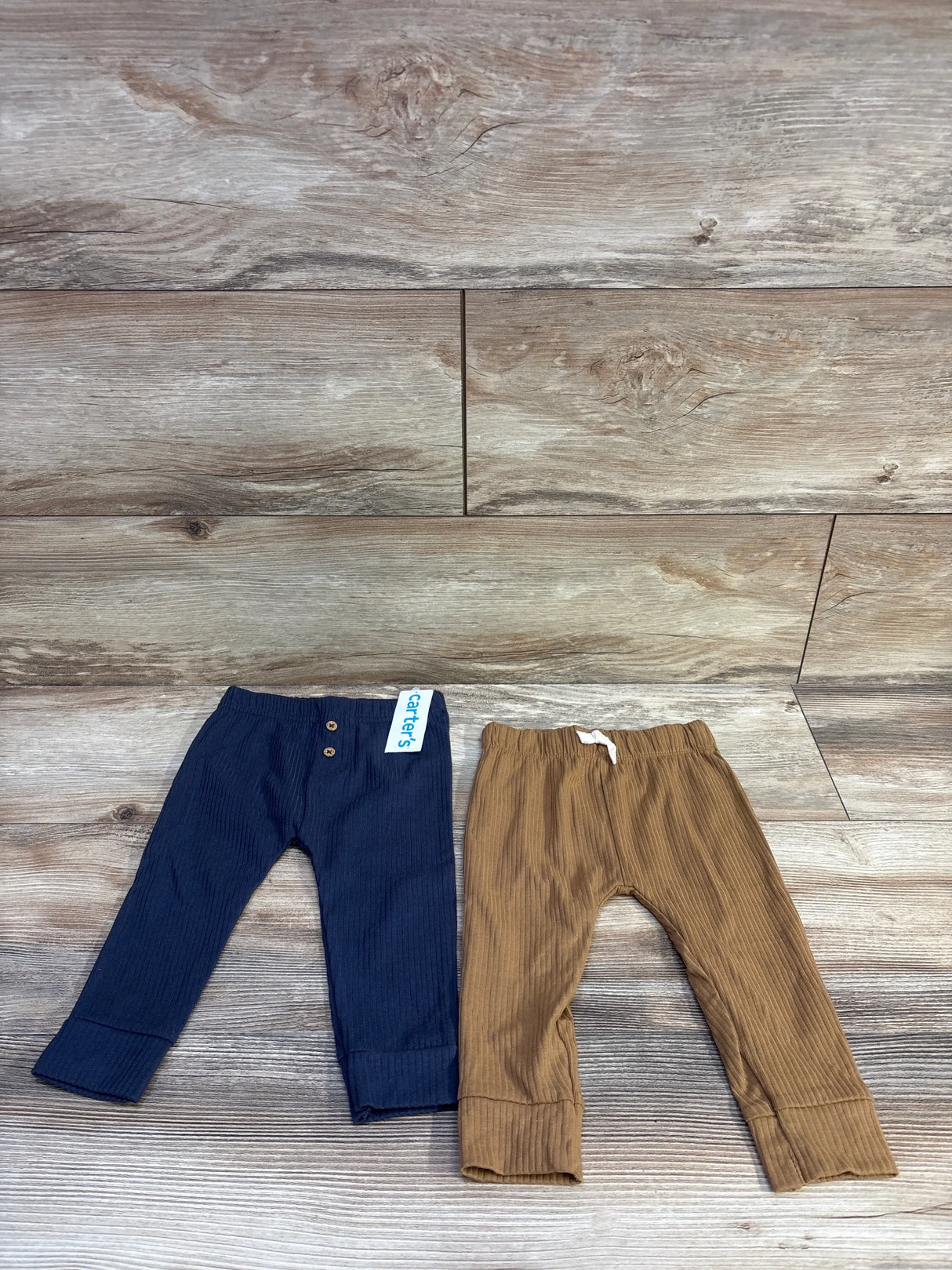 Top-down view of two Carters ribbed baby pants, navy and brown, showing fold-over cuffs and elastic waists on wood background