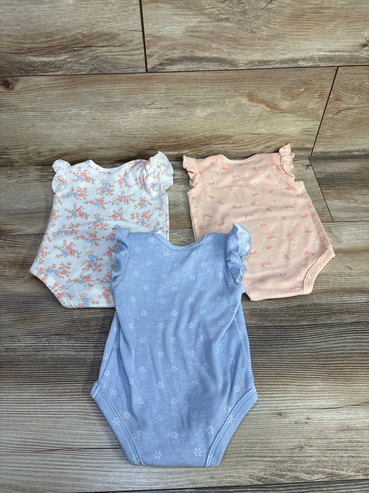 Top view of three Nicole Miller flutter-sleeve baby bodysuits (white floral, peach, blue) showcasing ruffled sleeves on wood background