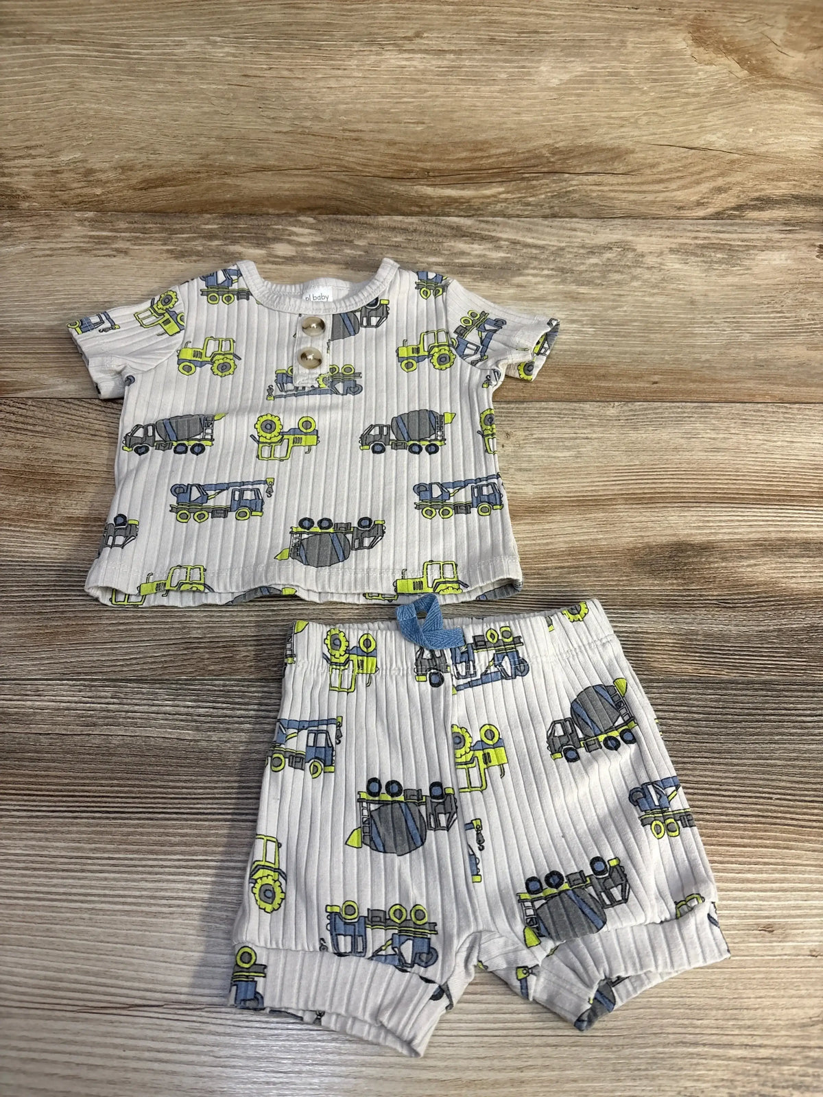 Flat lay of beige baby two-piece ribbed shirt and shorts with yellow-blue truck print, buttoned top and elastic waist on wood