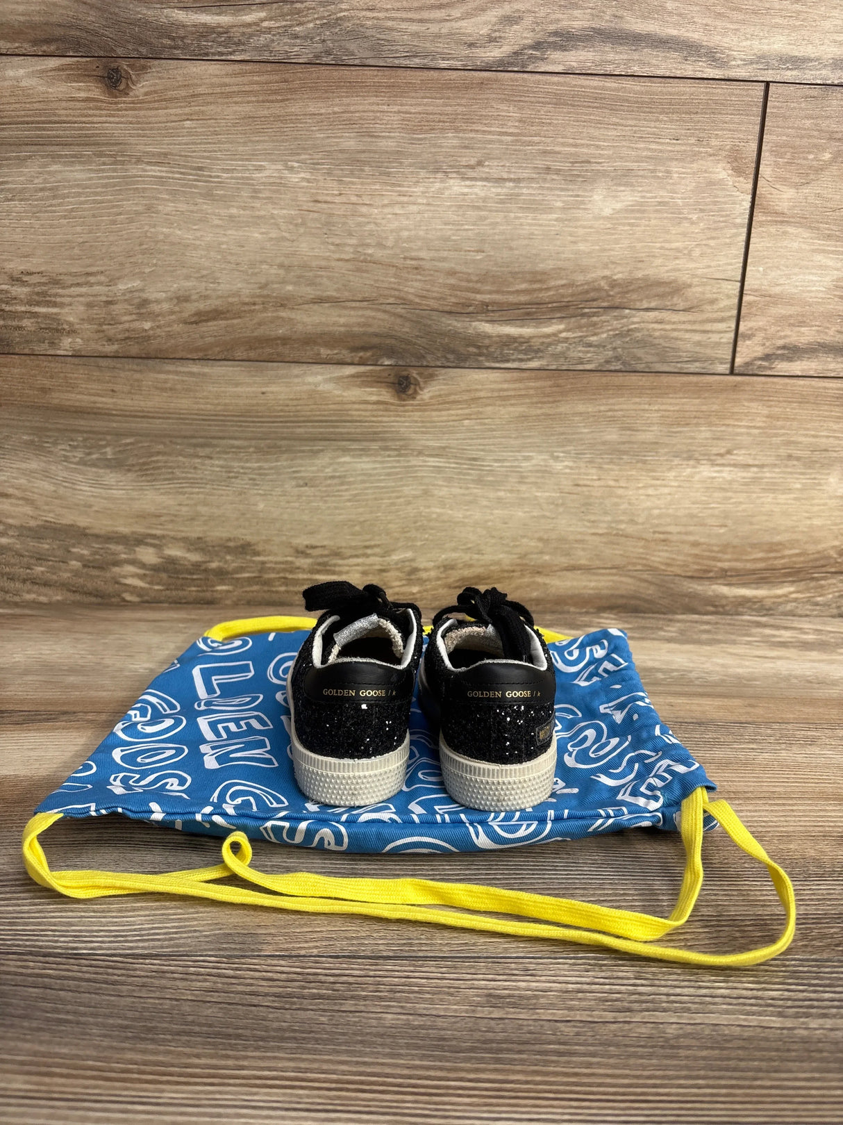 Back view of black glitter Golden Goose kids sneakers on blue branded drawstring bag with yellow straps on wooden floor