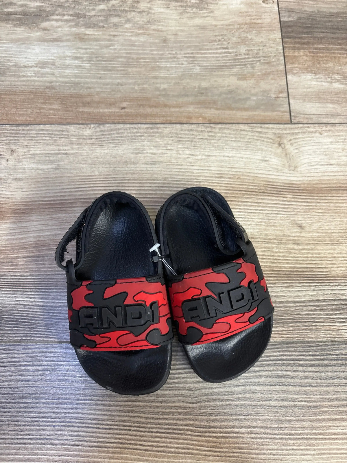 Top view of NWOT And1 Camo Slide Sandals Red sz 5-6c showcasing black slide with red and black camo strap and AND1 logo on wood plank floor