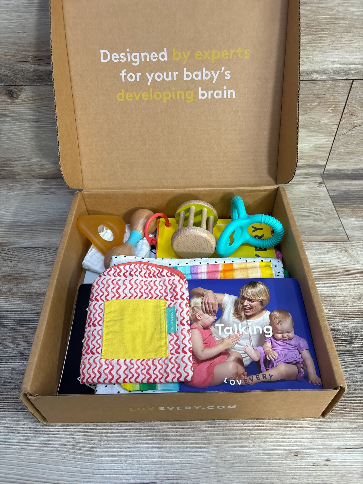 Top view of Lovevery The Charmer Play Kit with wooden rattle, silicone teether, framed mirror, soft book, and cloths on wood floor