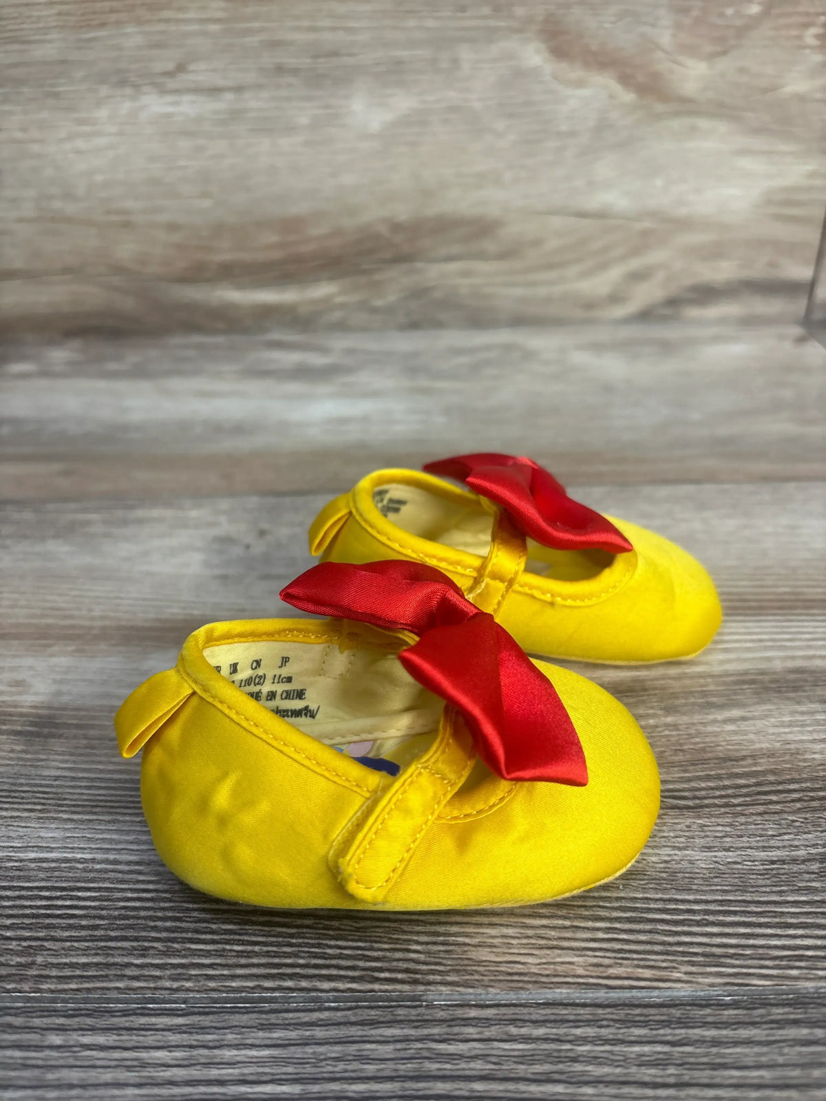 Angled view of Disney Baby Snow White soft sole shoes, size 1/2C, with red bows and pull tabs on a wood-look surface