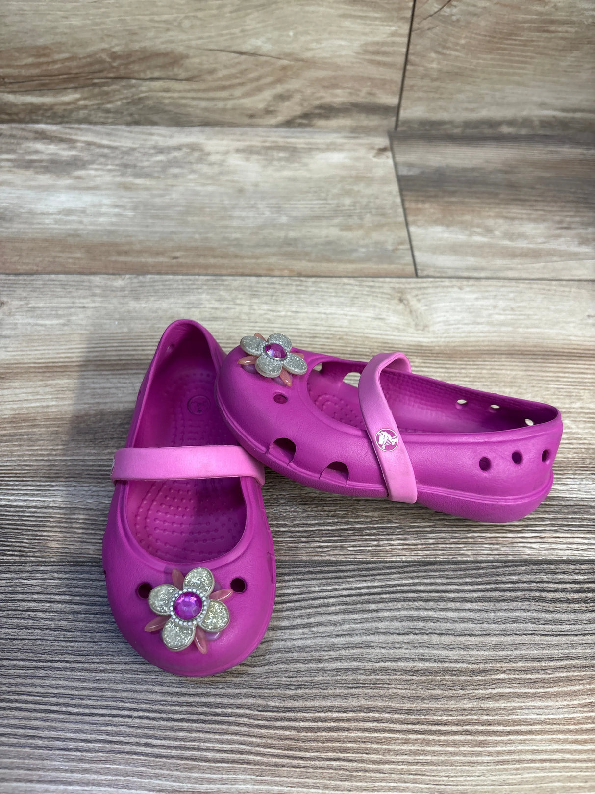 3/4 view of pink Crocs Keeley flats size 7C showing flower charm with purple gem and heel strap on wood floor