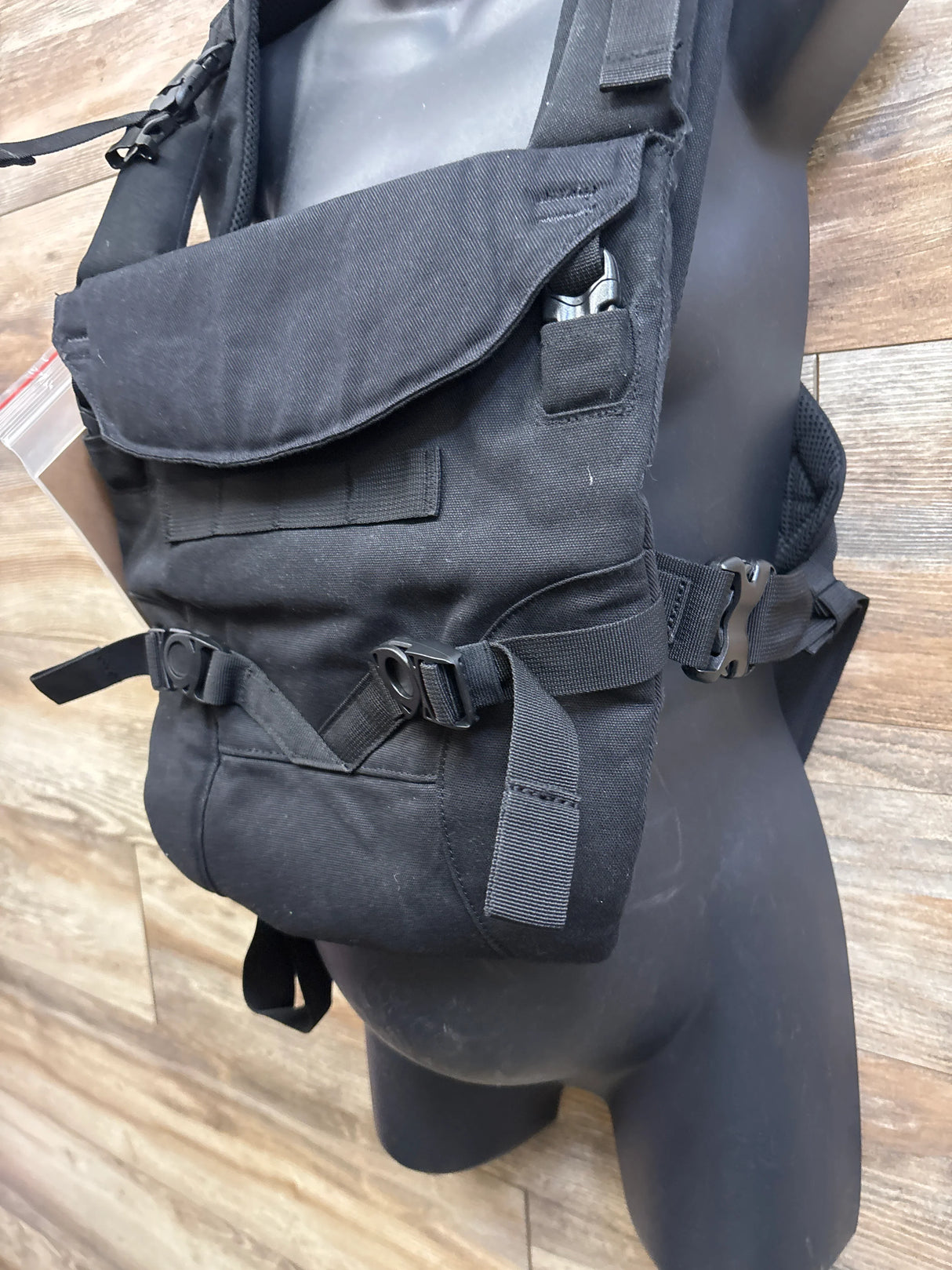 Side view of black Tactical Baby Gear carrier with front flap pocket and adjustable straps on mannequin, wood floor background