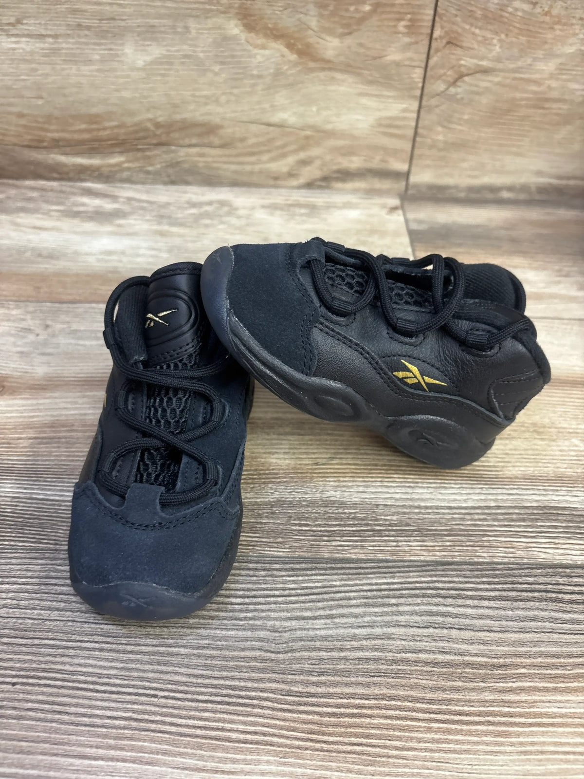 Top and side view of Reebok Question Mid Toddler Lux sneakers in black, showcasing gold logos on a wood floor