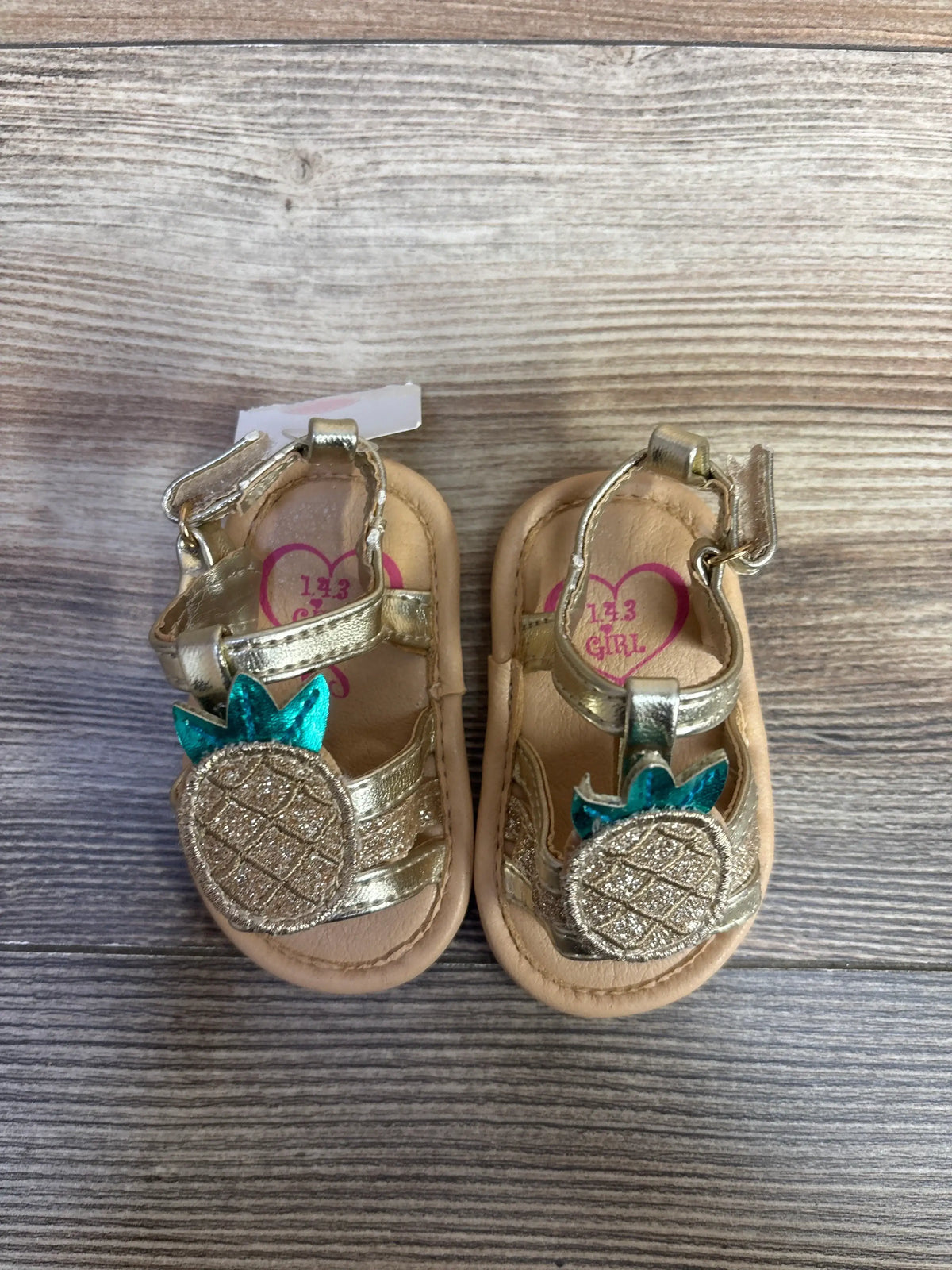 Top view of NEW 143 girls pineapple sandals size 1C with metallic pineapple appliques and hook and loop straps on wood grain