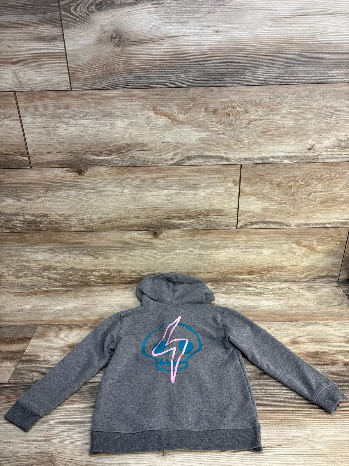 Gray kids hoodie with blue skull and pink lightning bolt graphic on back, laid on wood floor