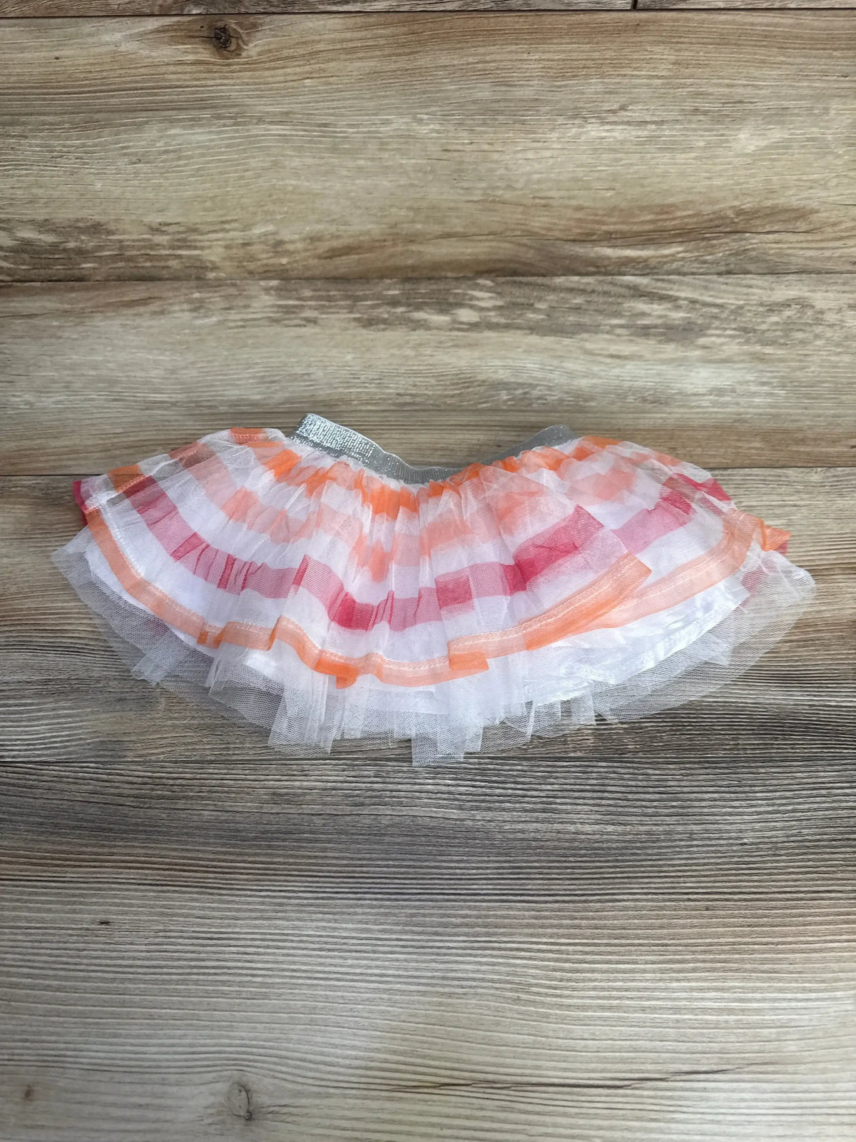 Flat lay of tutu skirt with layered white tulle, pink-orange bands and silver elastic waistband on wood, size 6 to 9 months