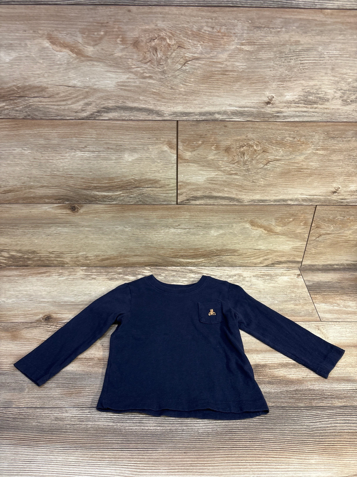 Front flat lay of Baby Gap navy long-sleeve pocket shirt showcasing a small embroidered tan bear on the chest pocket with wood plank background