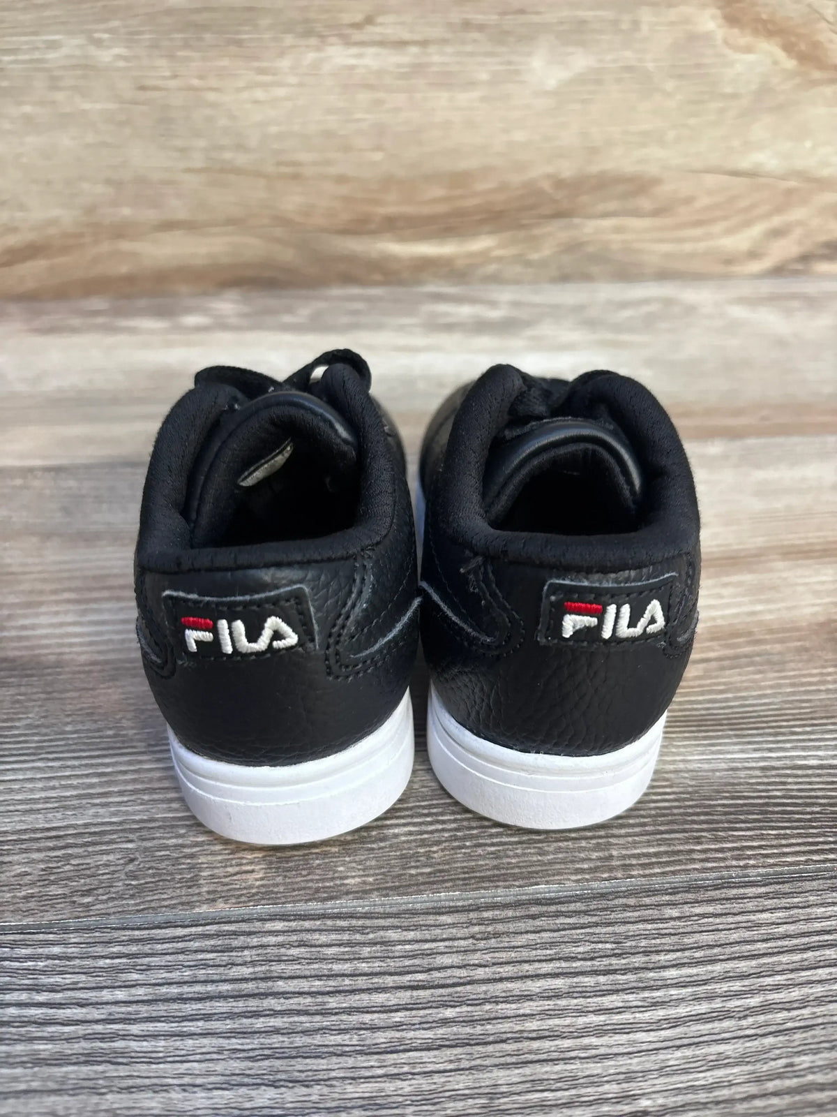 Pair of black FILA sneakers with white soles on wooden floor, rear view showing logo