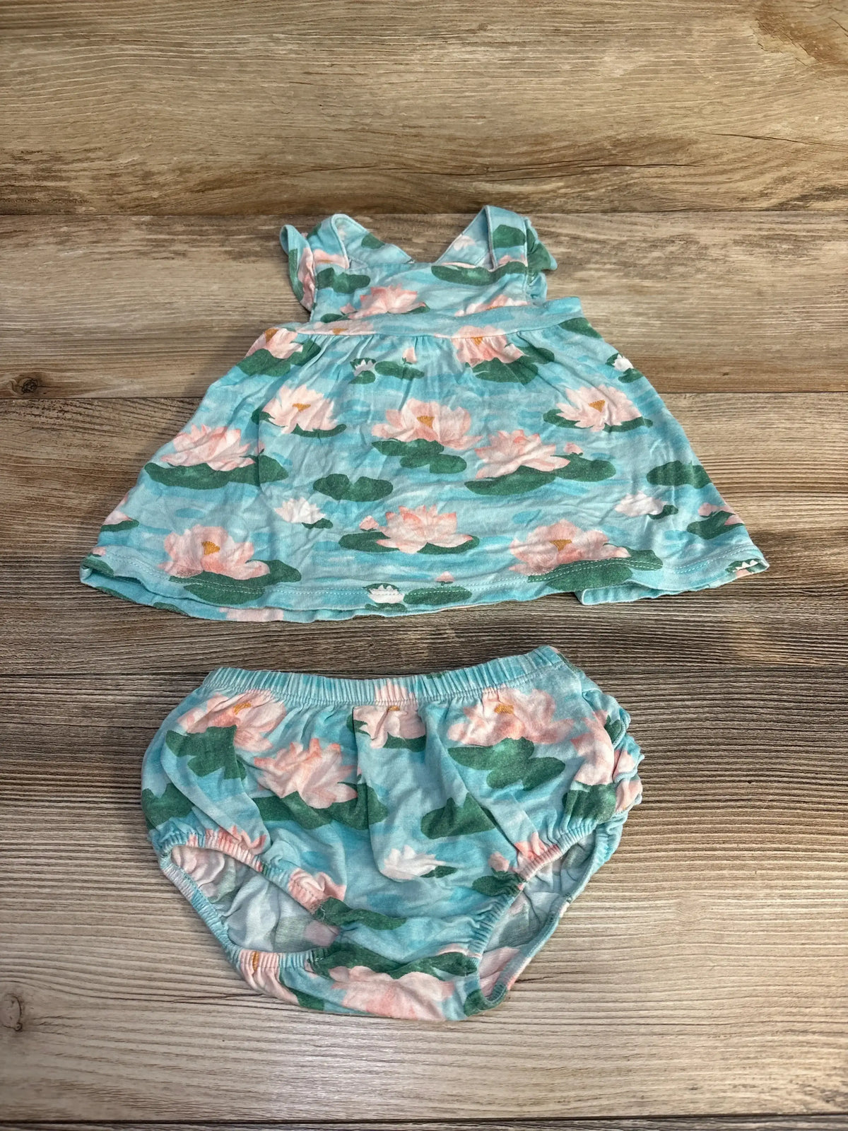 Flat lay of 2-piece pinafore and bloomers in aqua lily pad print with ruffle straps and elastic waist on wood background