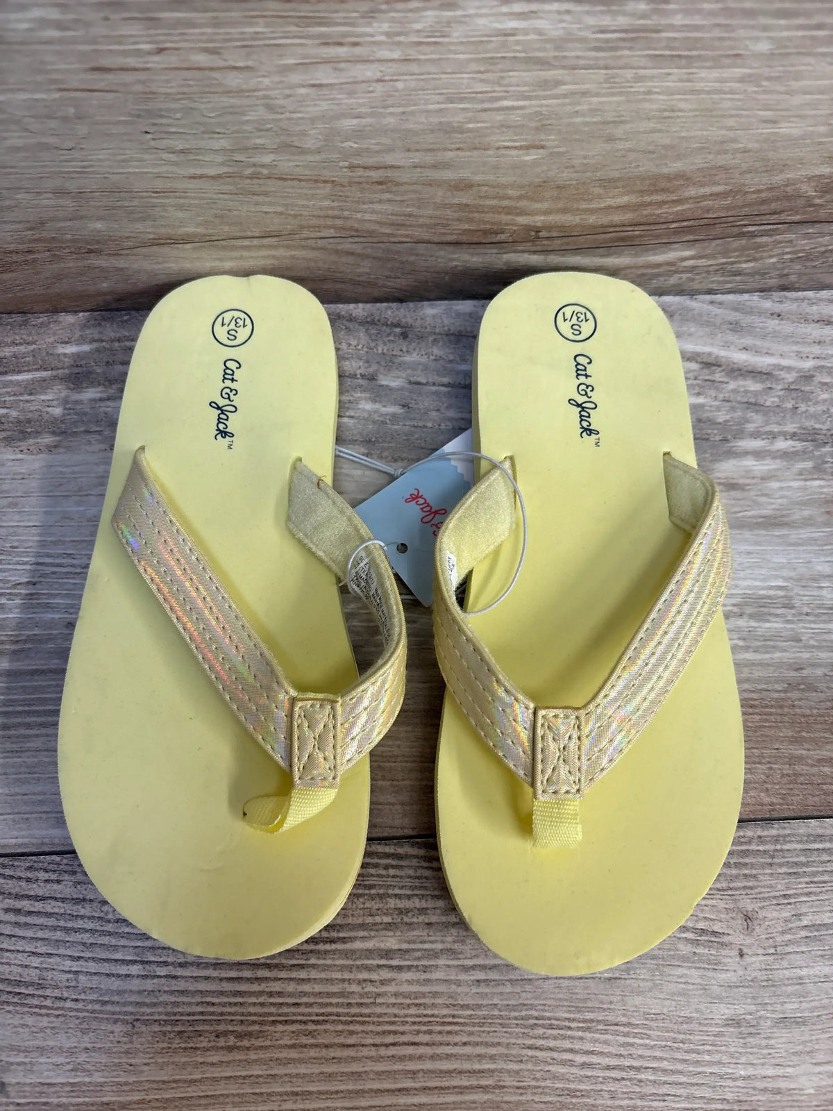 Top view of Cat & Jack Ava flip-flops in yellow size 13C/1Y showing iridescent straps on a wood floor