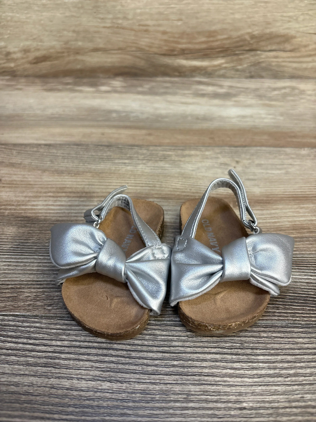 Front view of Old Navy toddler sandals showcasing metallic silver faux-leather tie bows and cork footbeds on wood background