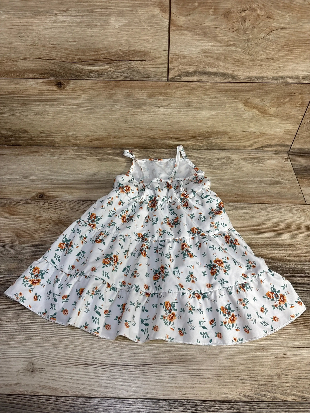 Front view of white tiered baby dress, orange floral print, thin straps and ruffled tiers on wood background
