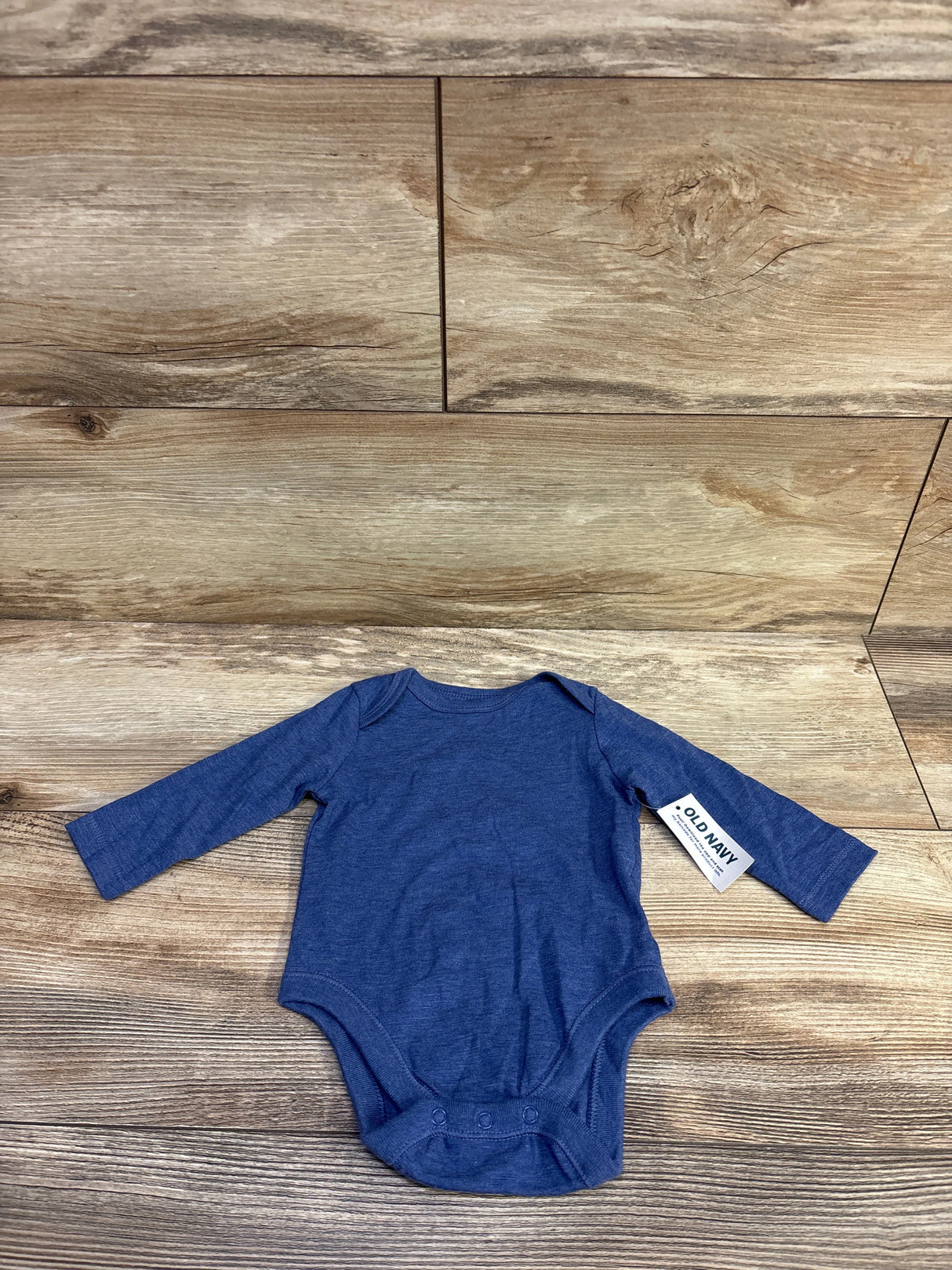 Flat-lay front view of Old Navy blue long-sleeve baby bodysuit size 0-3 months showing crew neck and three-snap crotch on wood background