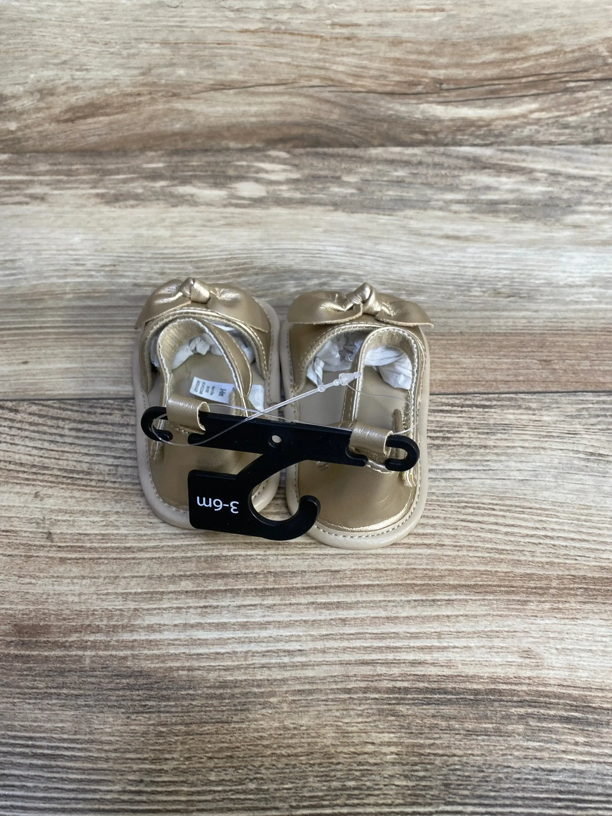 Top-down view of Child of Mine gold infant sandals showing bows, soft padded soles, and size 3-6M hanger on wood plank background