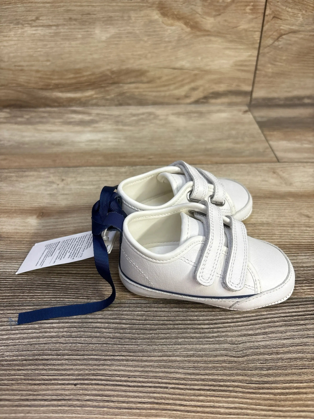 Angled view of a pair of white Janie & Jack toddler sneakers, size 3/4C, showcasing double straps on a wood-grain floor
