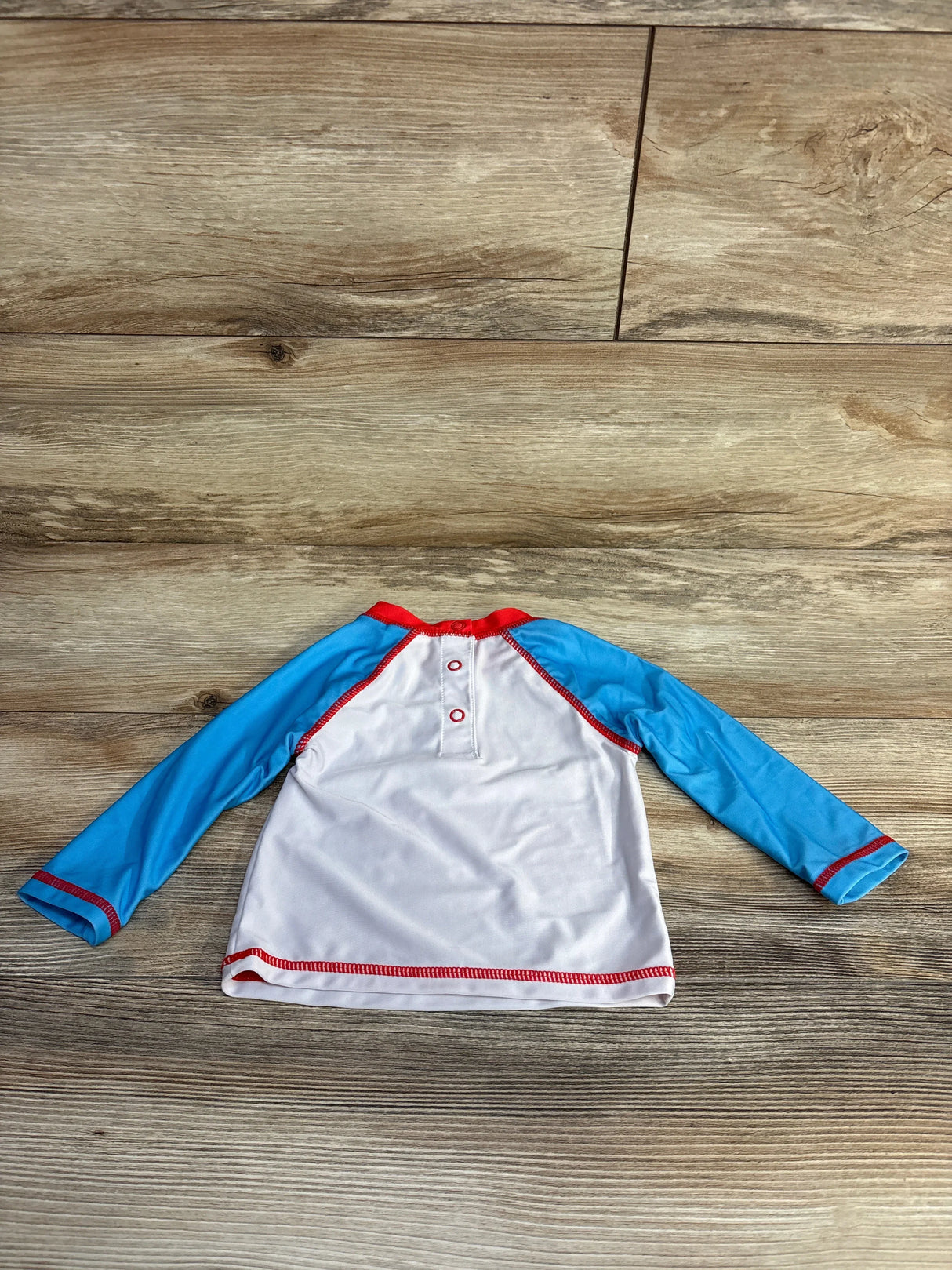 Back view of baby rashguard with white body, blue long sleeves, red trim and two back snap buttons on wood floor background