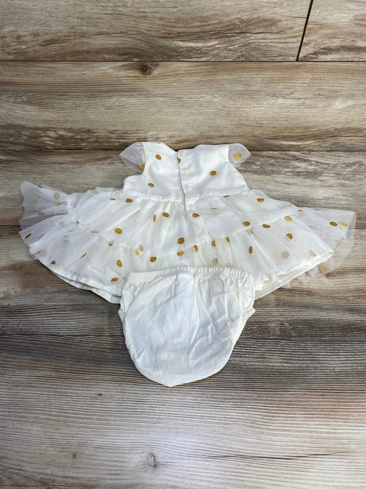 Flat lay of cream baby dress with matching bloomers showcasing gold polka dot tulle overlay and back button closure on wood background