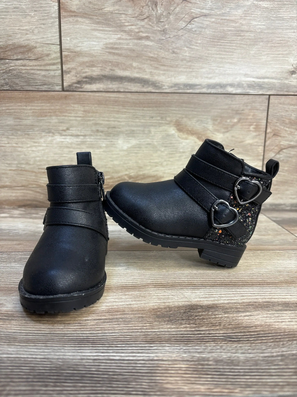 Black toddler boots with glitter heart buckles, side zipper, and sturdy sole on wood floor
