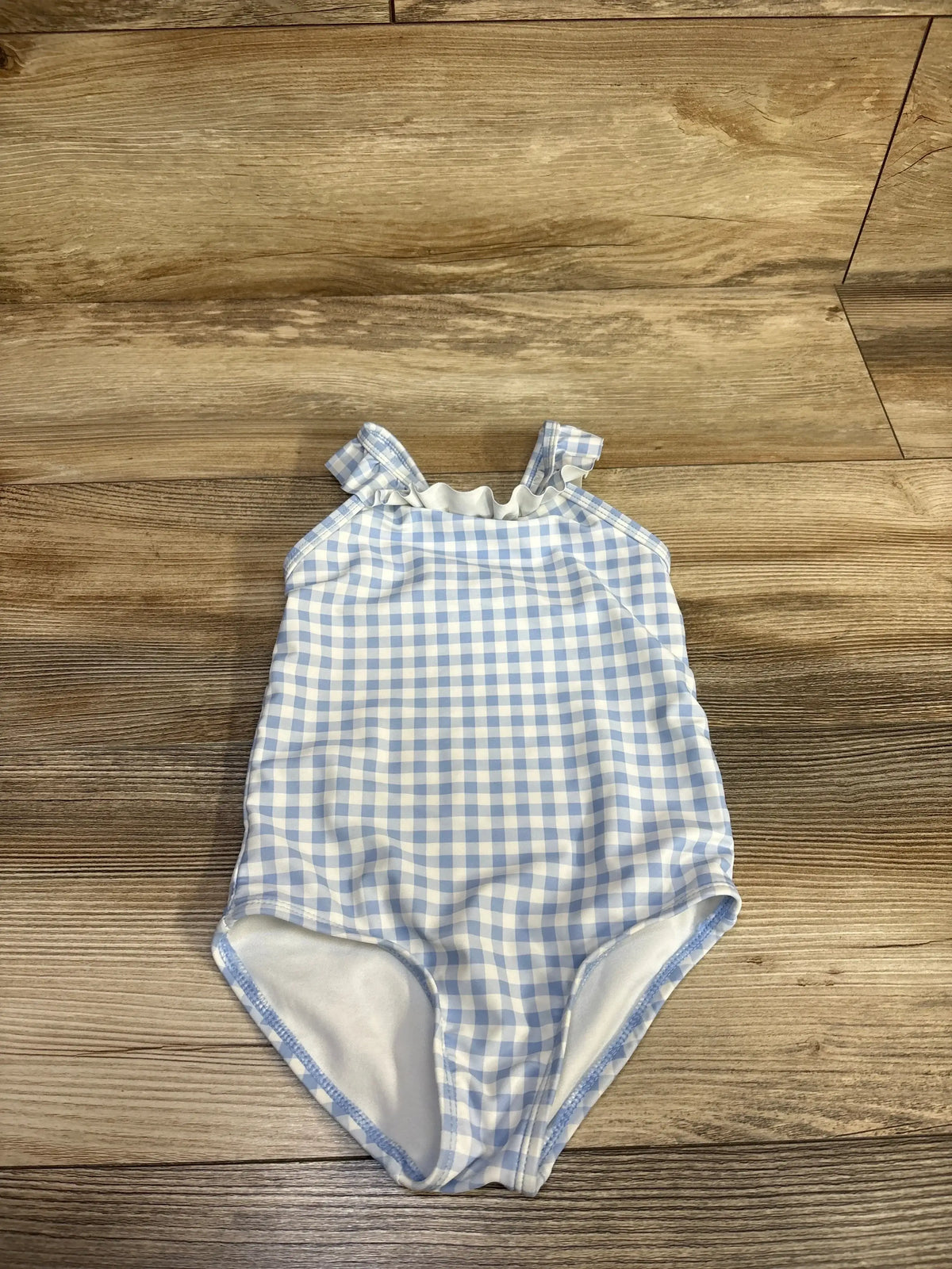 Flat lay of toddler one-piece blue and white gingham swimsuit with ruffle shoulder straps on wood floor, size 2T