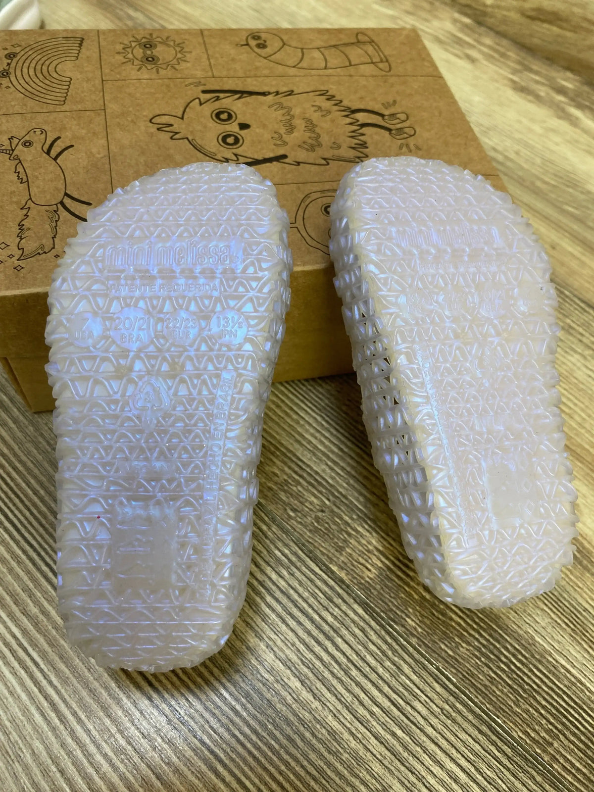 Soles-up view of Mini Melissa Campana Papel jelly flats showing textured translucent soles on wood floor with illustrated shoe box