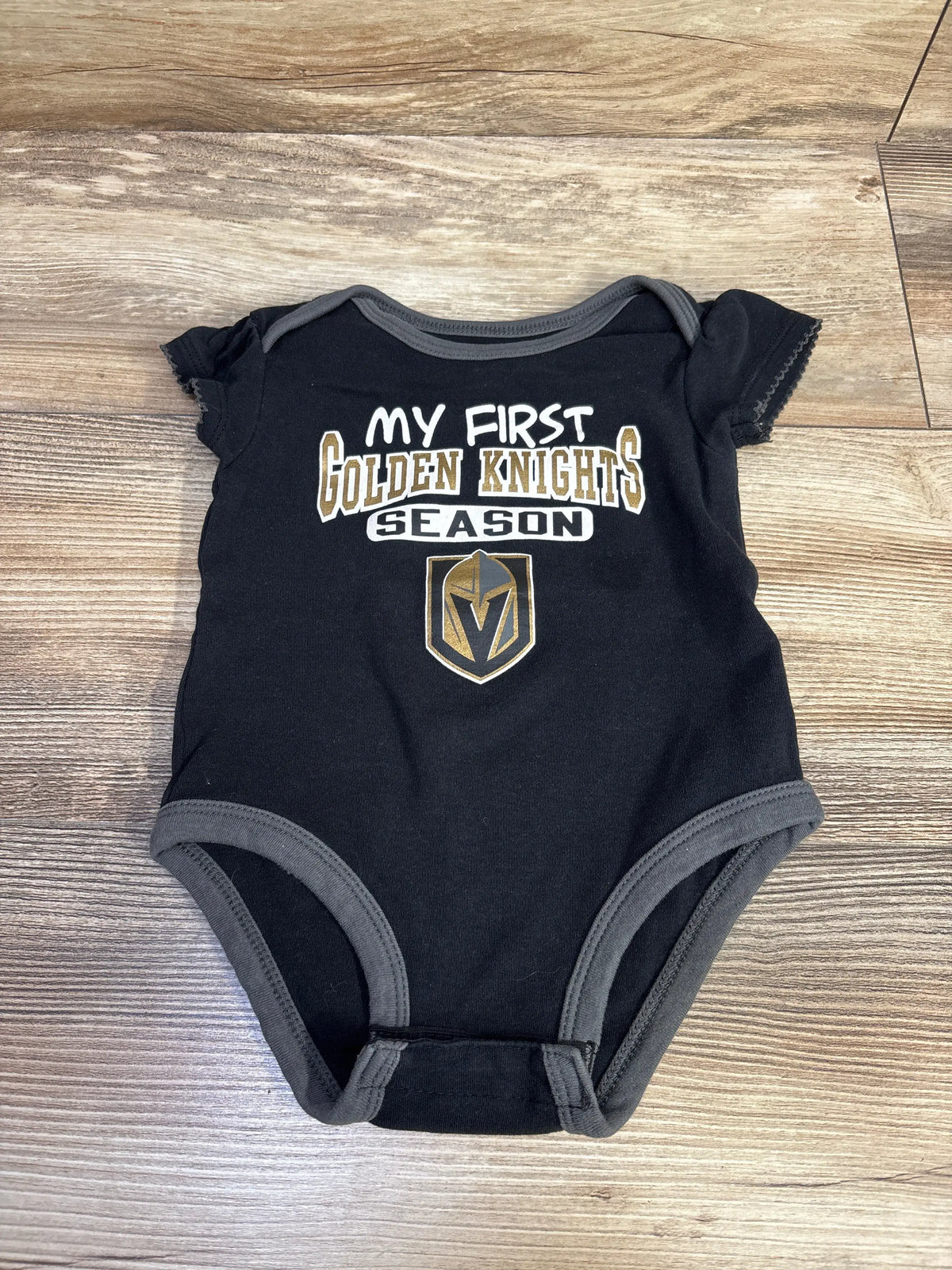 Front view of black NHL My First Golden Knights Season bodysuit showcasing team graphic and snap closures on a wood floor