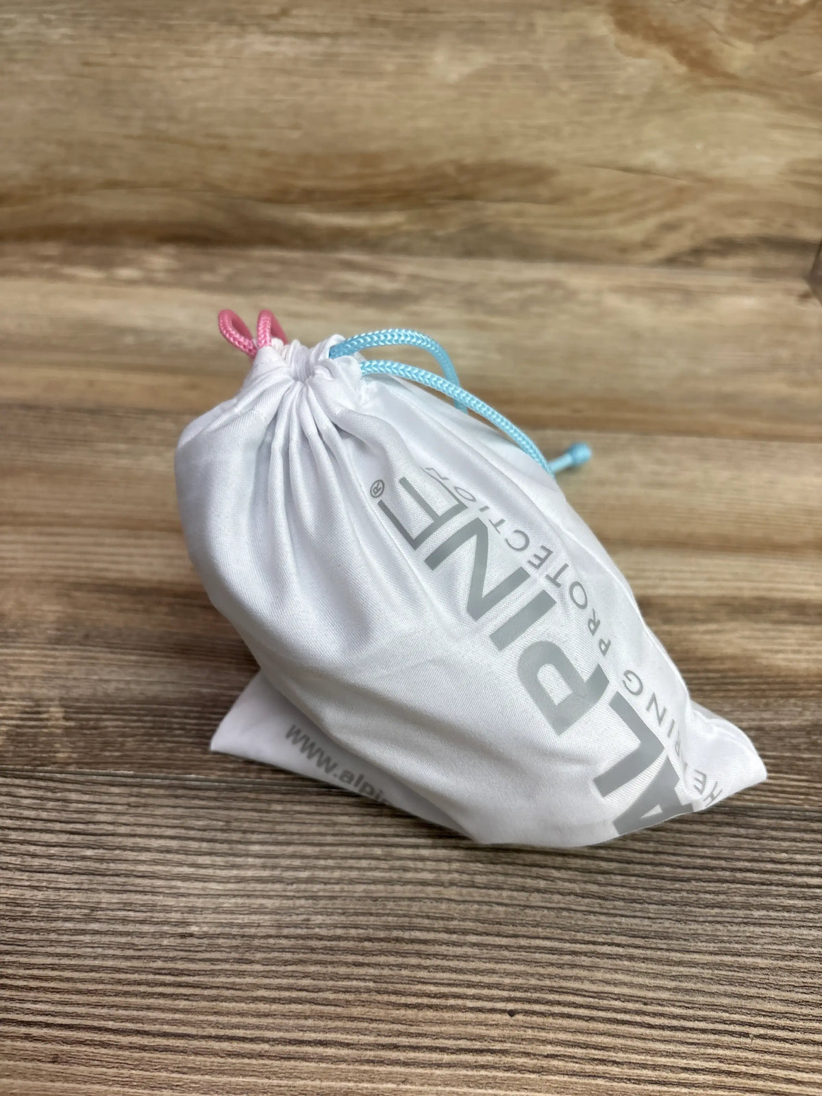 Angled view of white Alpine Muffy Baby ear muffs pouch showcasing pink and blue drawstrings on a wood-grain surface