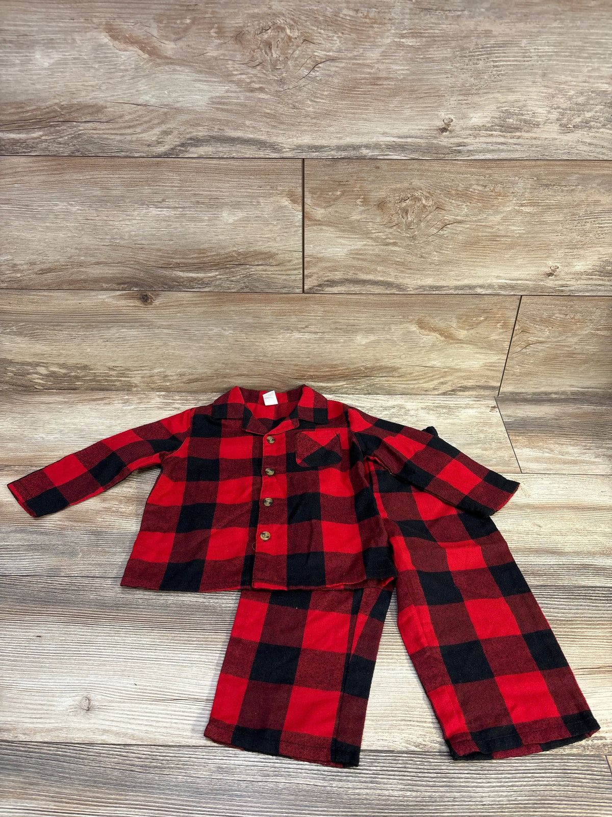Flat lay of red and black plaid toddler coat-style pajama set with button-front top and matching pants on wood floor