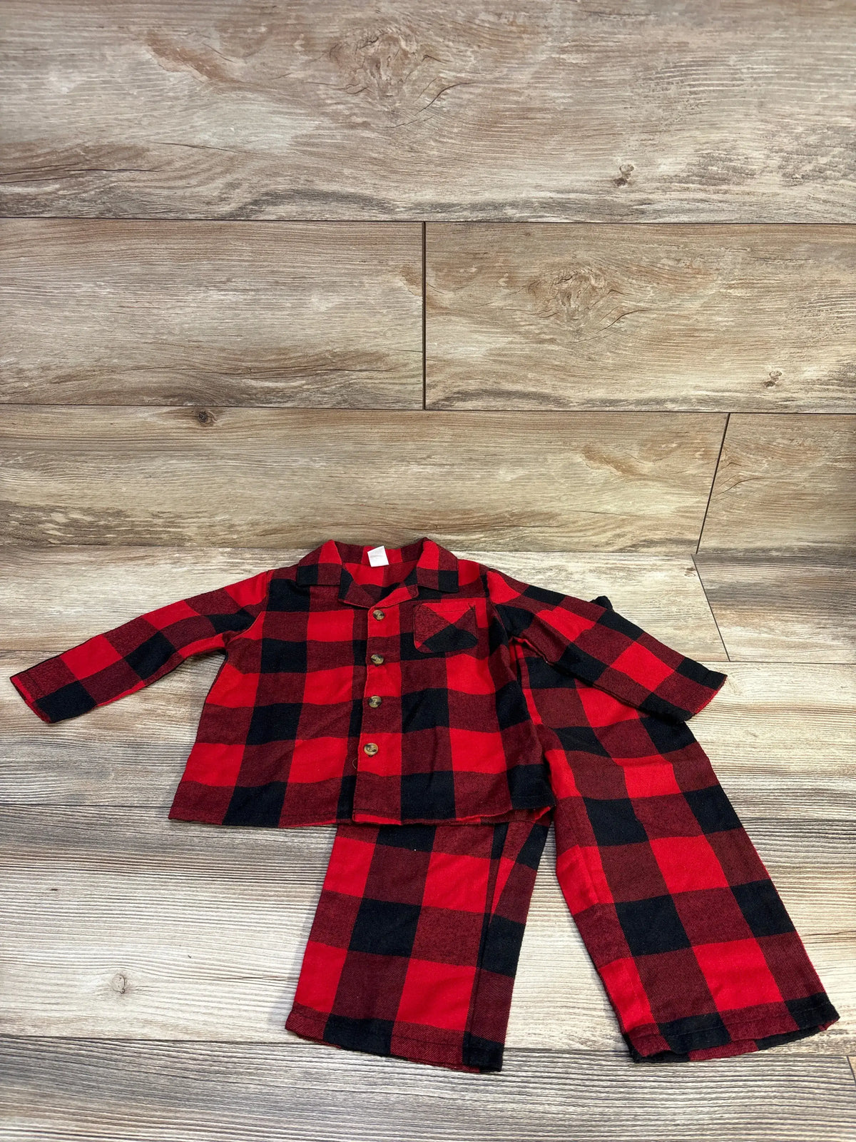 Flat lay of red and black plaid toddler coat-style pajama set with button-front top and matching pants on wood floor
