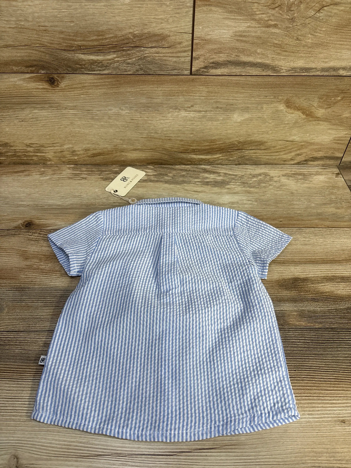 Back view of light blue and white striped seersucker baby button-up shirt showing short sleeves, gathered yoke, and brand tag on wooden background