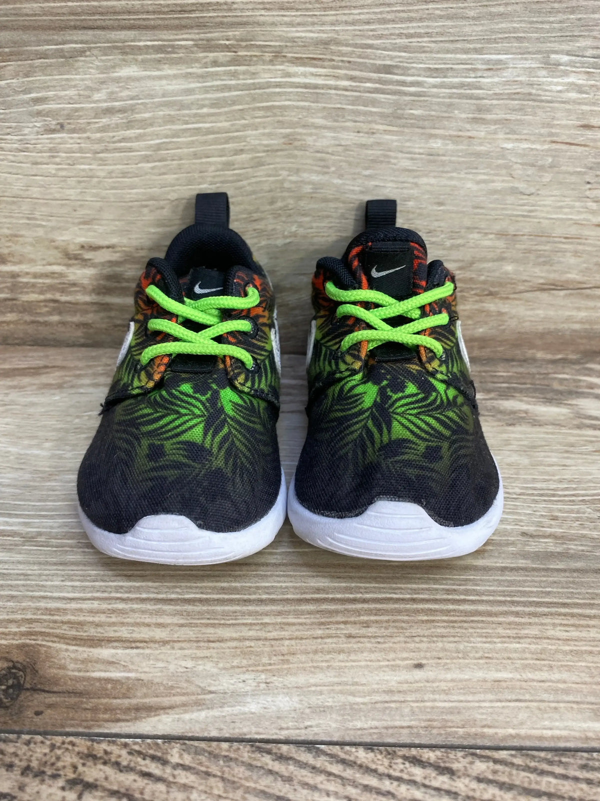 Front view of Nike Roshe Run toddler sneakers, black and green leaf print, neon green laces, white sole on wood background
