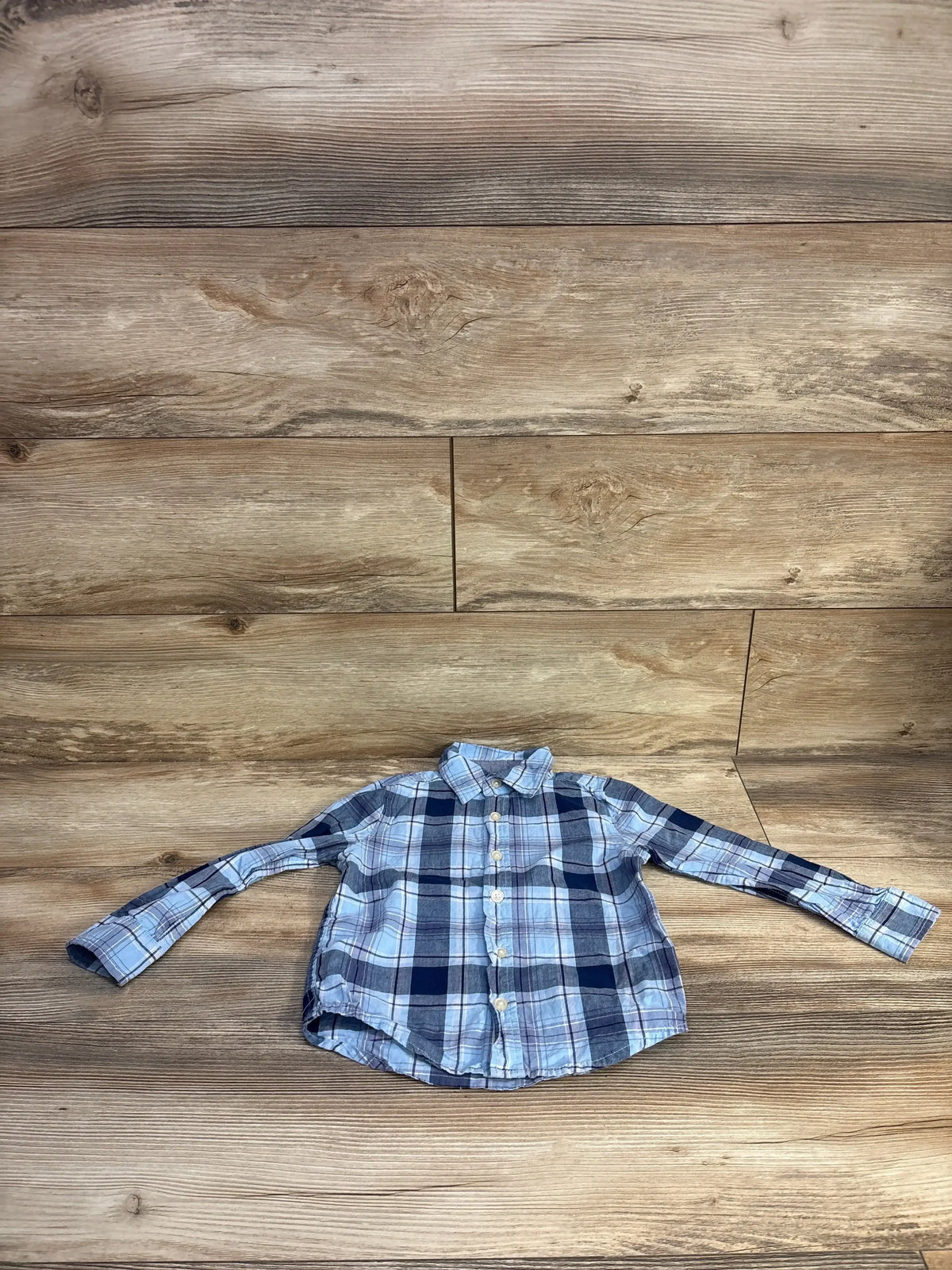 Front view of boys long sleeve blue plaid OshKosh button up shirt showing buttons and collar on wooden plank background, size 3T