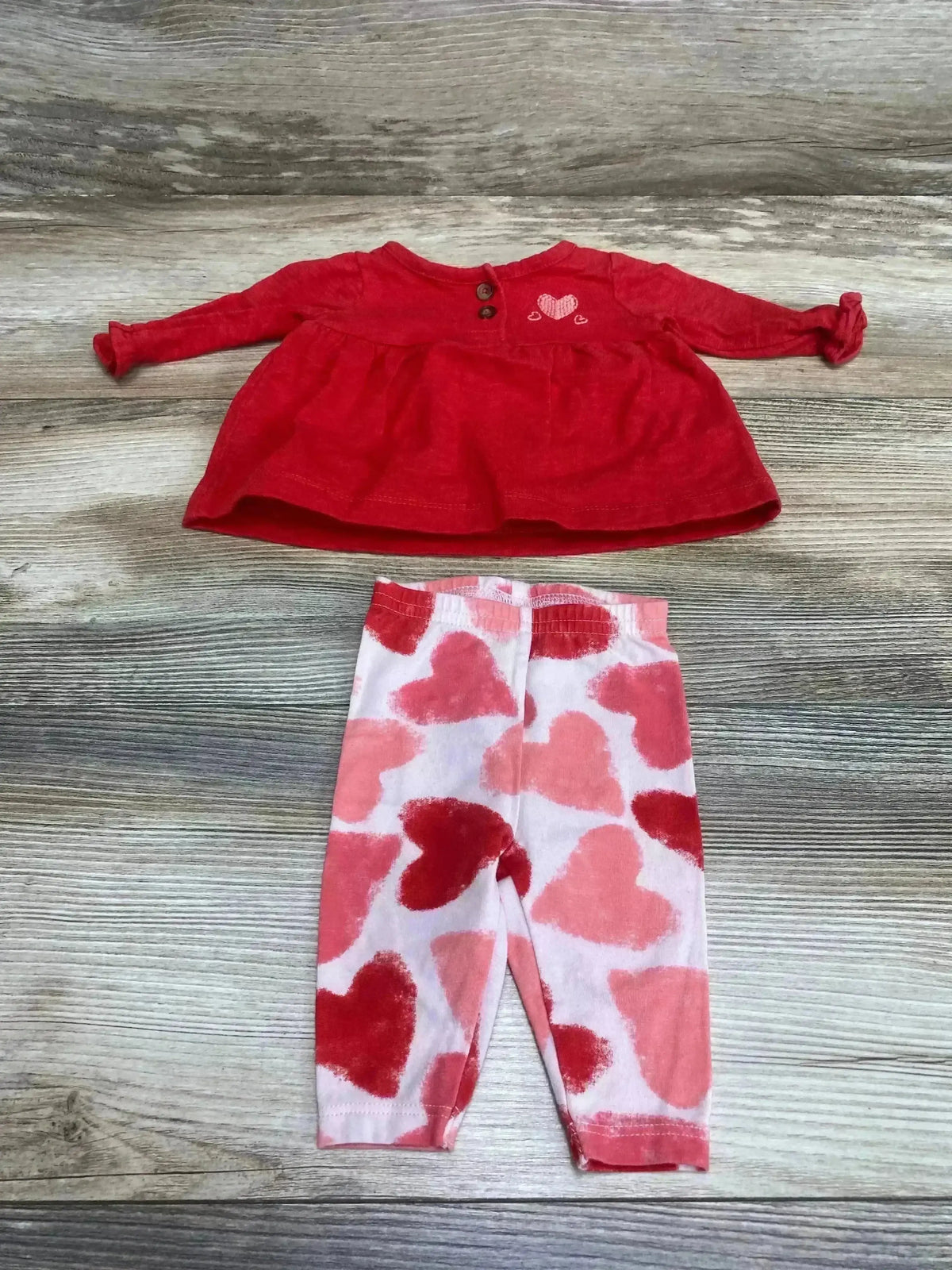 Flat lay of newborn red Henley top with buttons and embroidered heart, and pink leggings with red hearts on wood