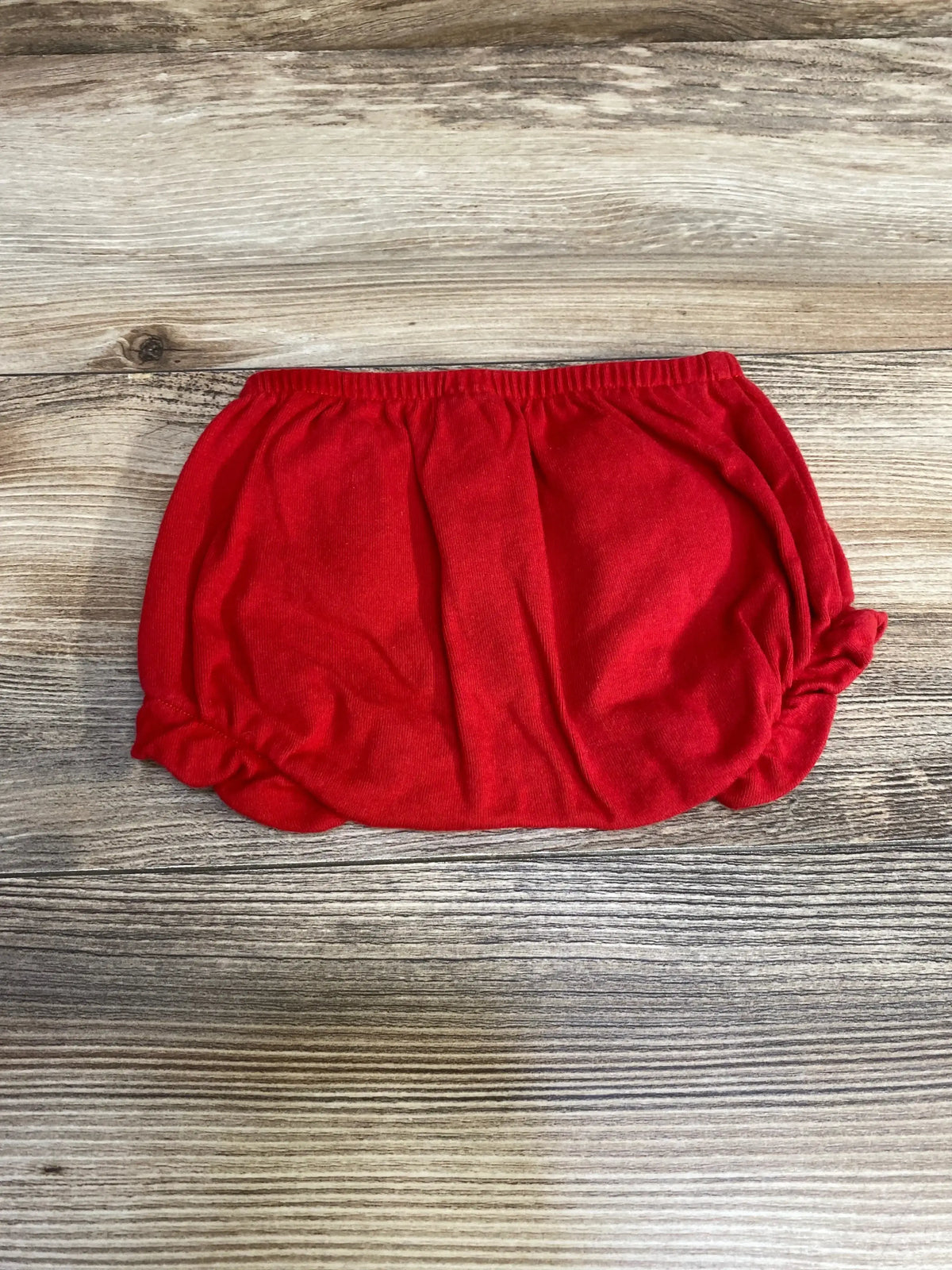 Top down view of red baby bloomers size 3 to 6 months with elastic waistband and gathered leg openings on wood background