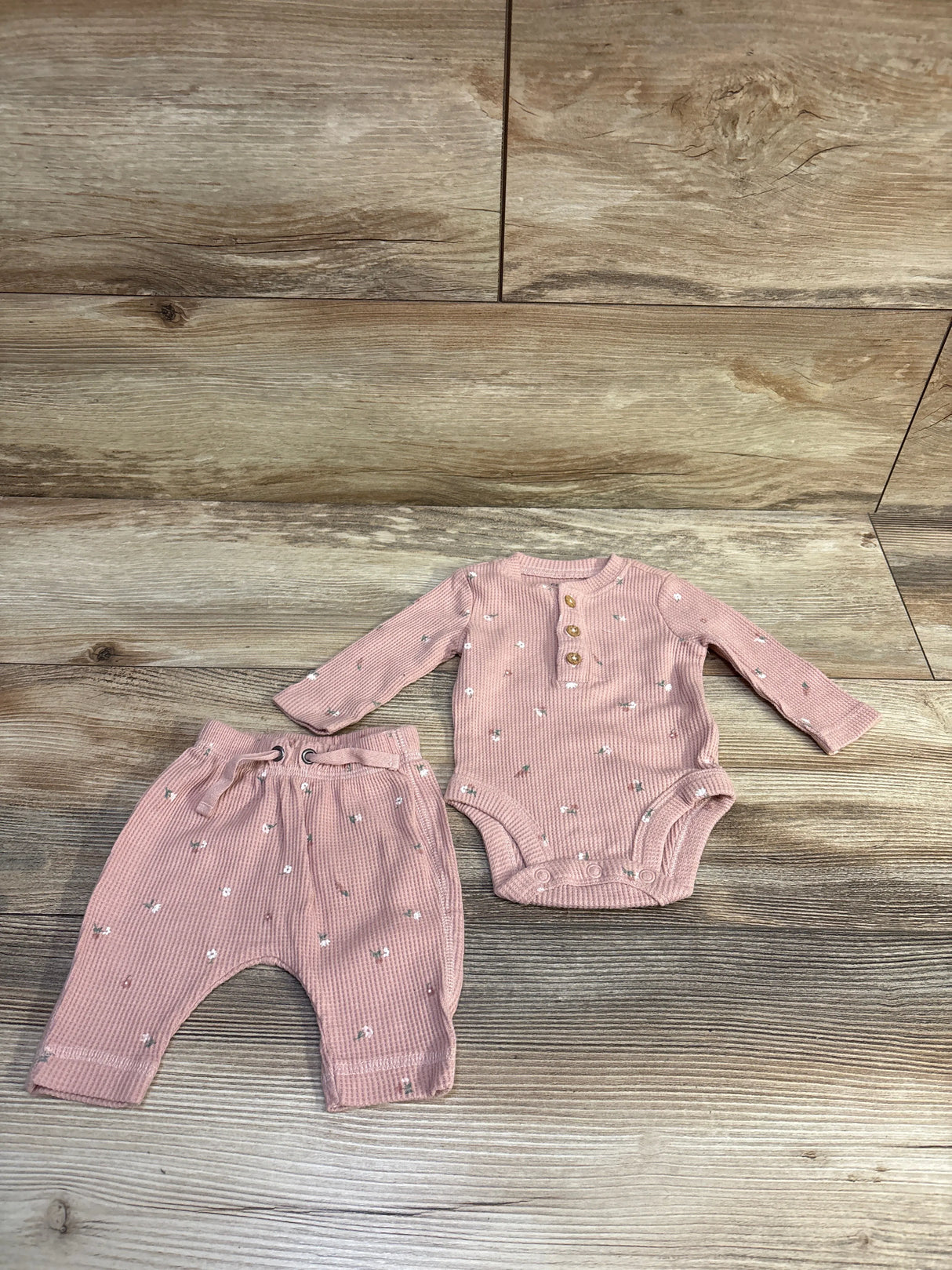 Top down view of newborn pink waffle knit Henley bodysuit and pants with wood buttons and small white flowers on wood floor
